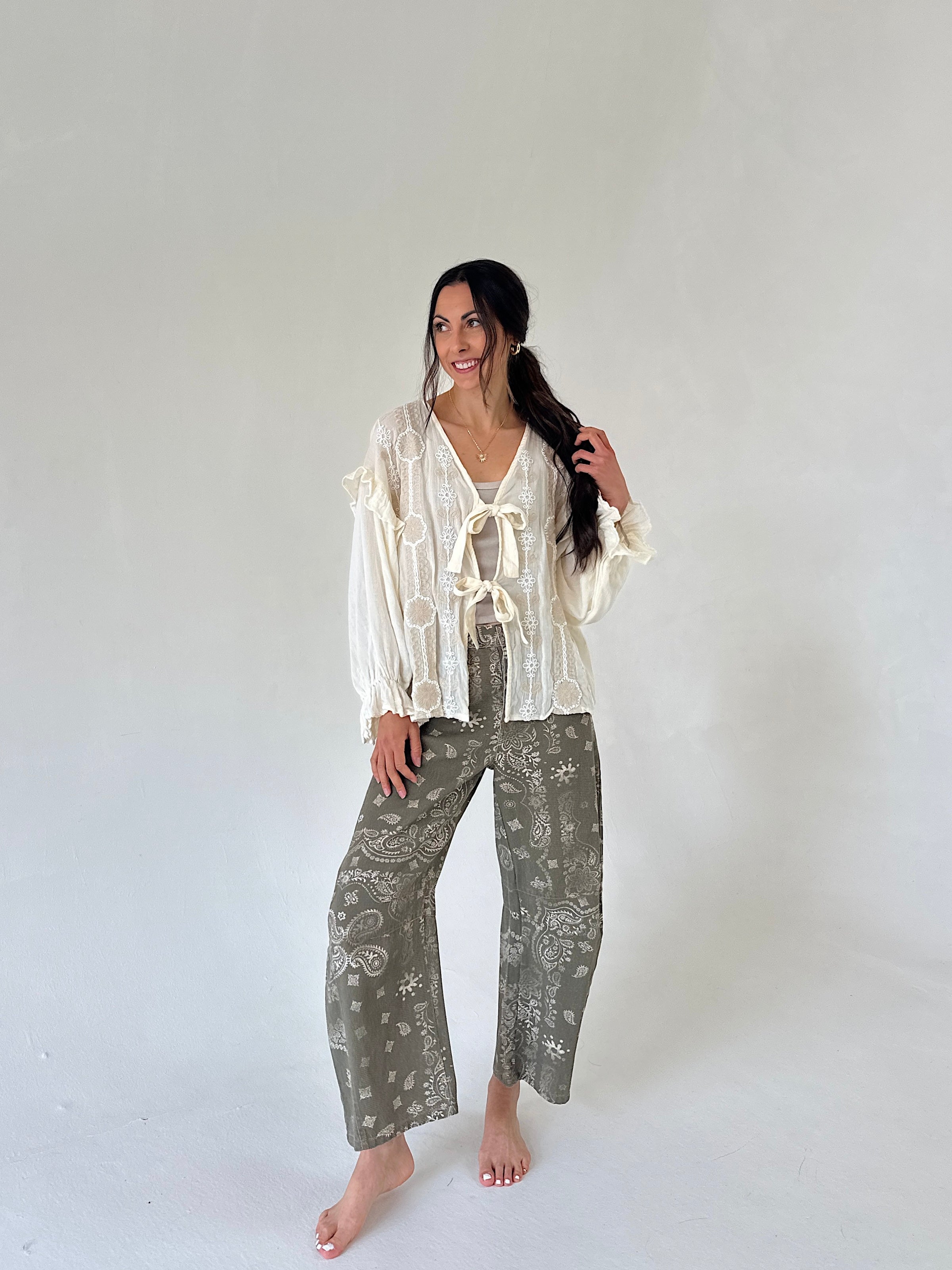 A woman modeling a bohemian-style outfit of a cream ruffled long-sleeve top and olive green patterned wide-leg pants, standing against a plain light gray studio background.