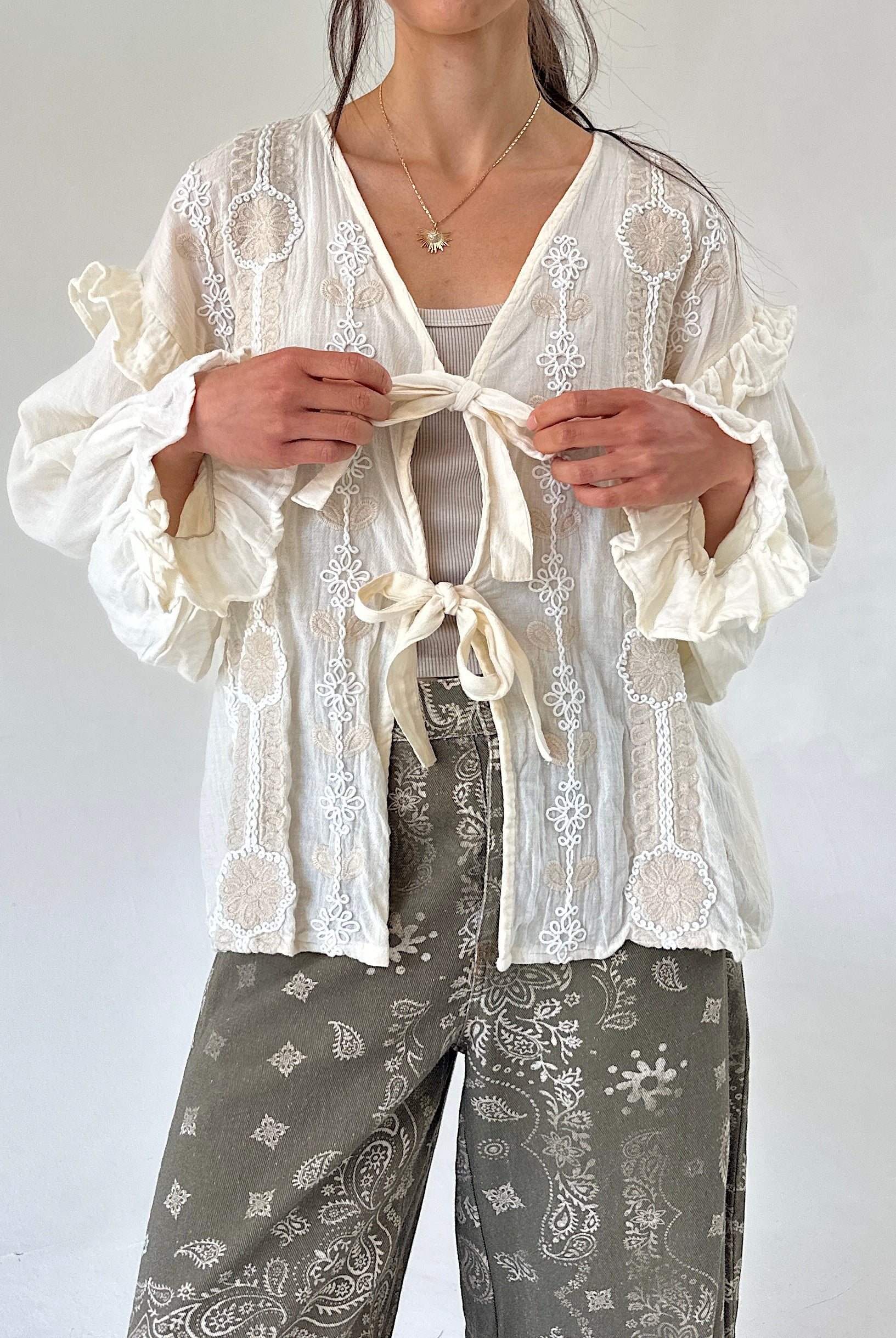 A woman wearing a cream tie-front long sleeve top with ruffled sleeves over a light gray top and gray paisley pants, photographed against a plain light background.