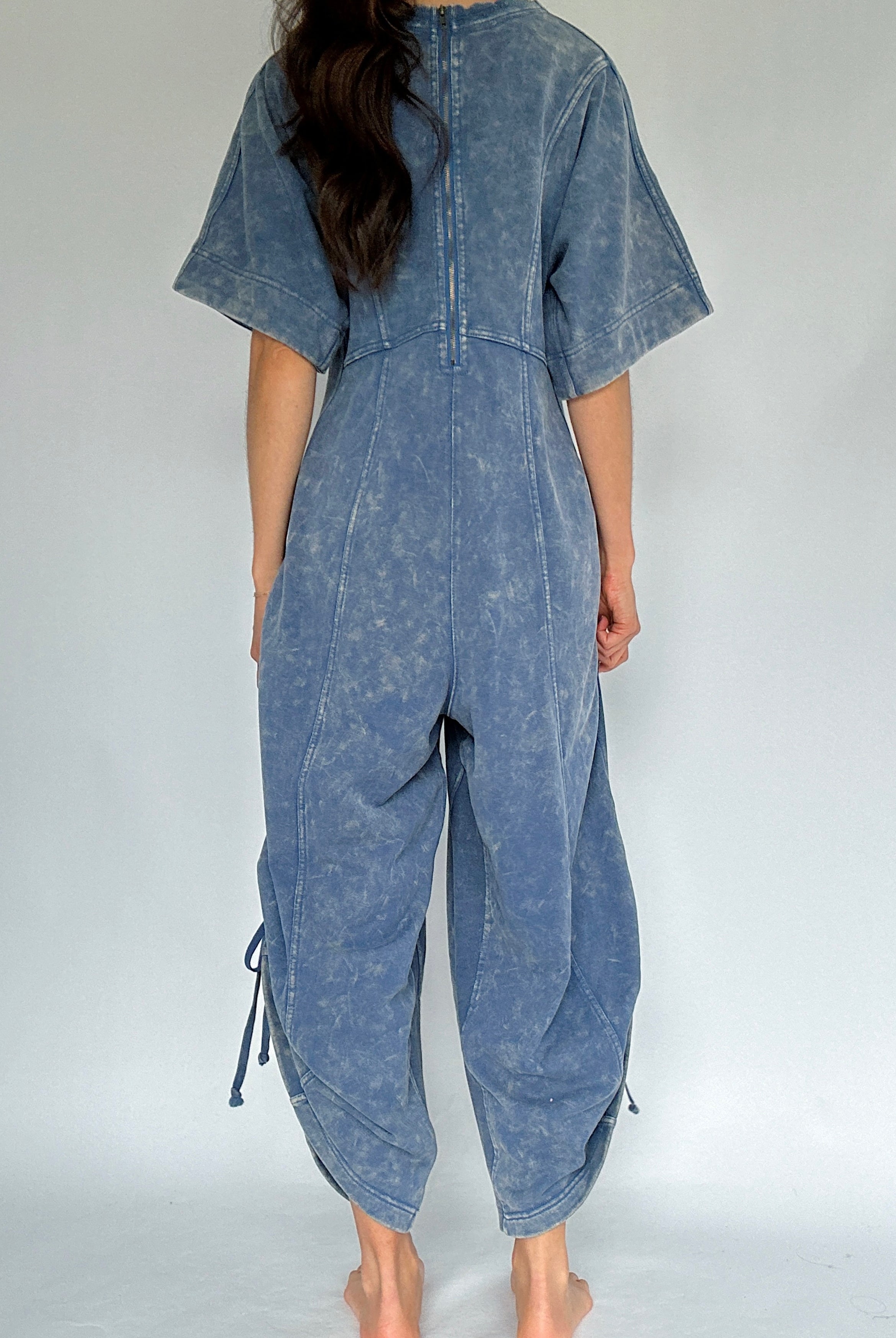 Person wearing a blue mineral-wash jumpsuit on a white background to show back view