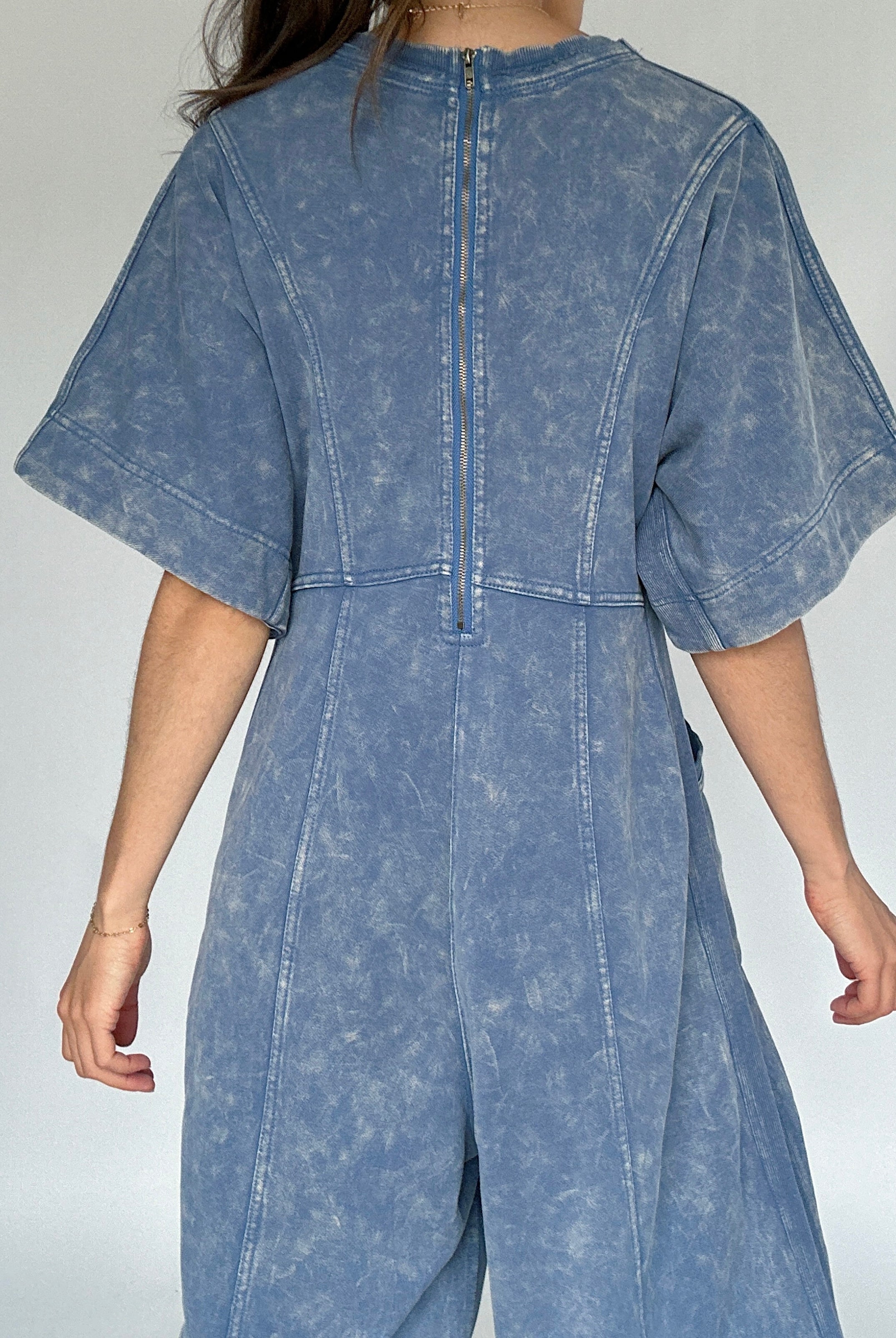 Person wearing a blue mineral-wash jumpsuit against a plain background to show back zip up detail