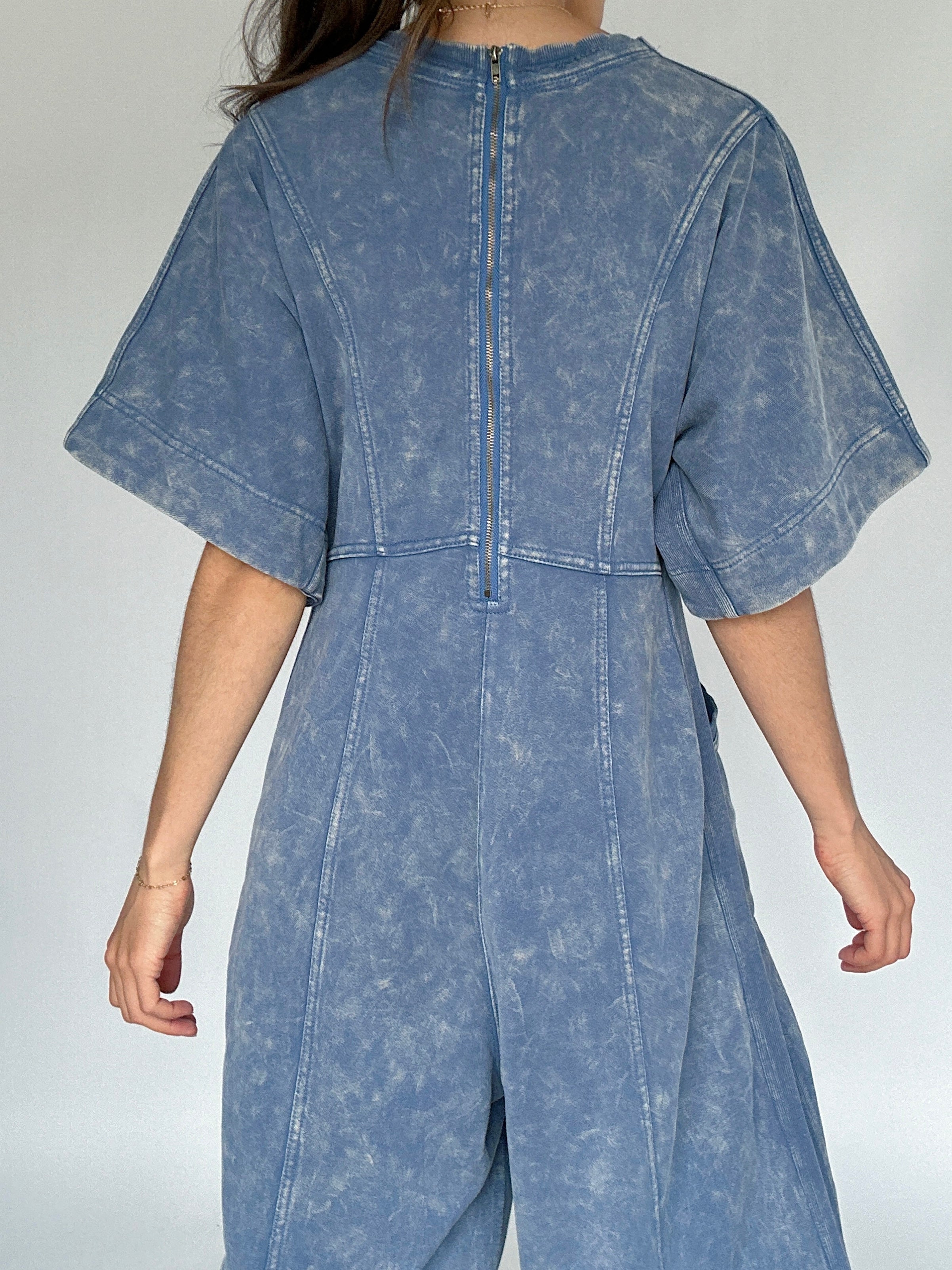Person wearing a blue mineral-wash jumpsuit against a plain background to show back zip up detail