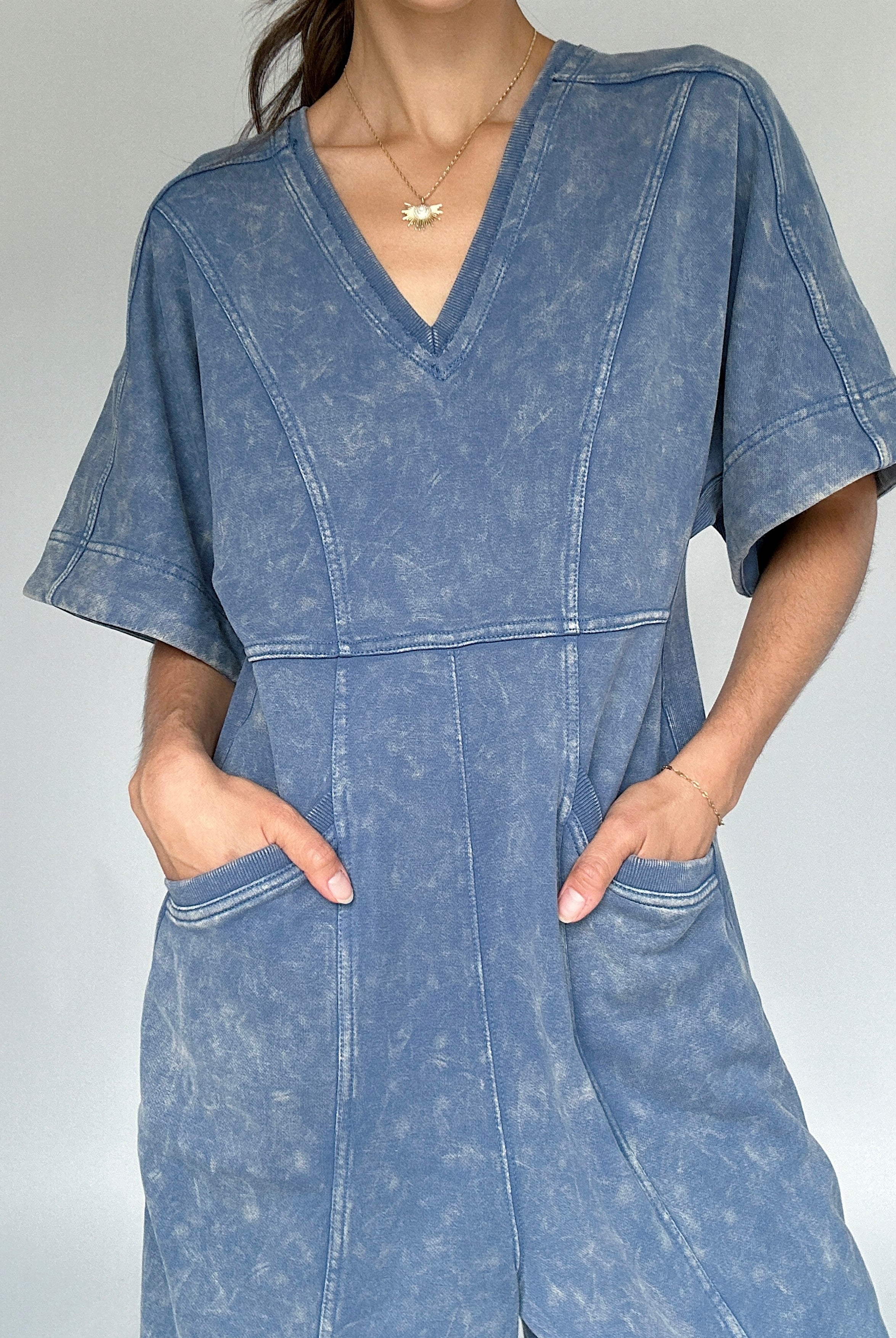 Person wearing a blue mineral-wash jumpsuit against a plain background up close to show v-neckline and pockets