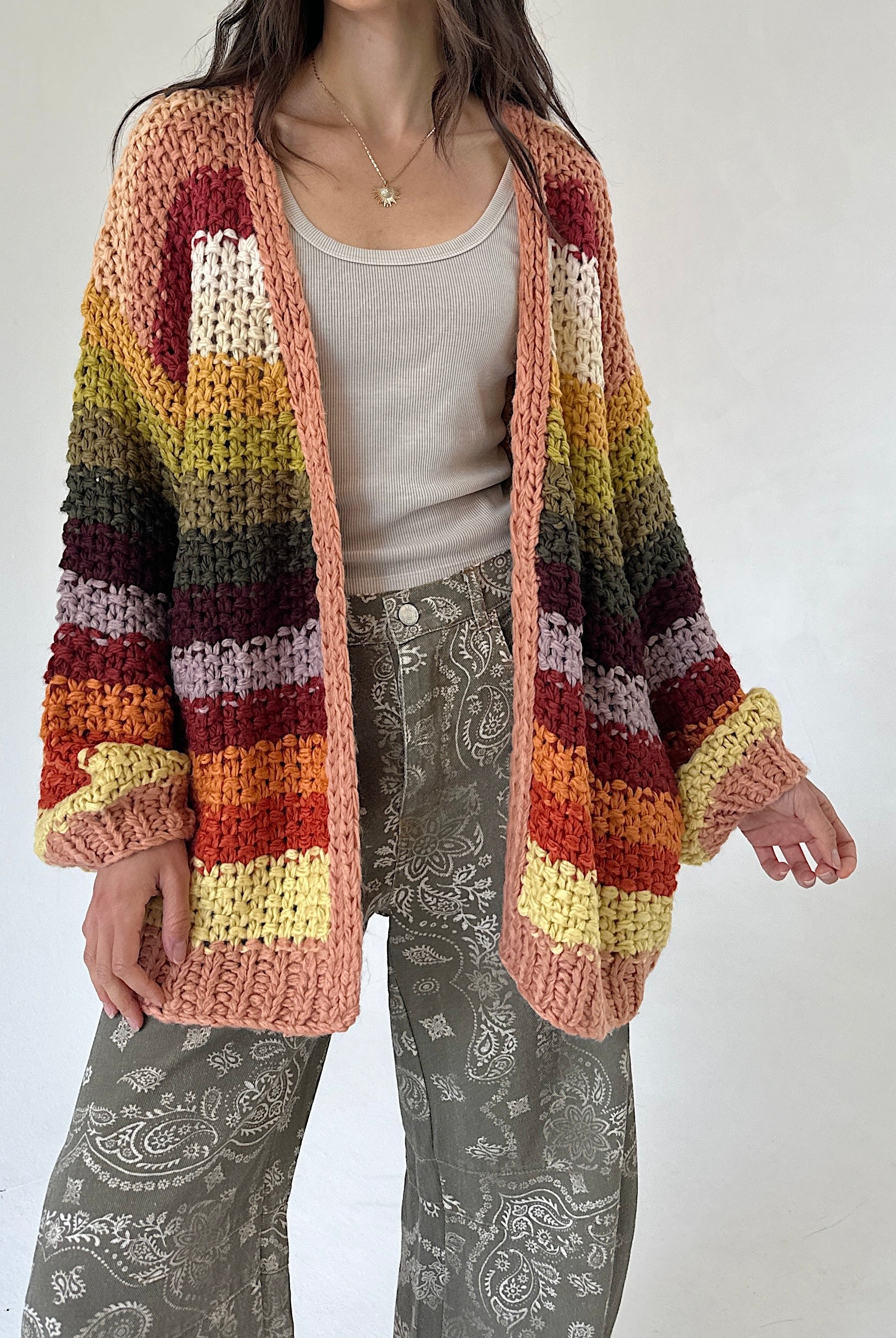 A person wearing a chunky knit open-front cardigan with horizontal multicolor stripes in peach, rust, orange, yellow, olive, and dark brown, layered over a light gray top and gray paisley pants against a plain light background.