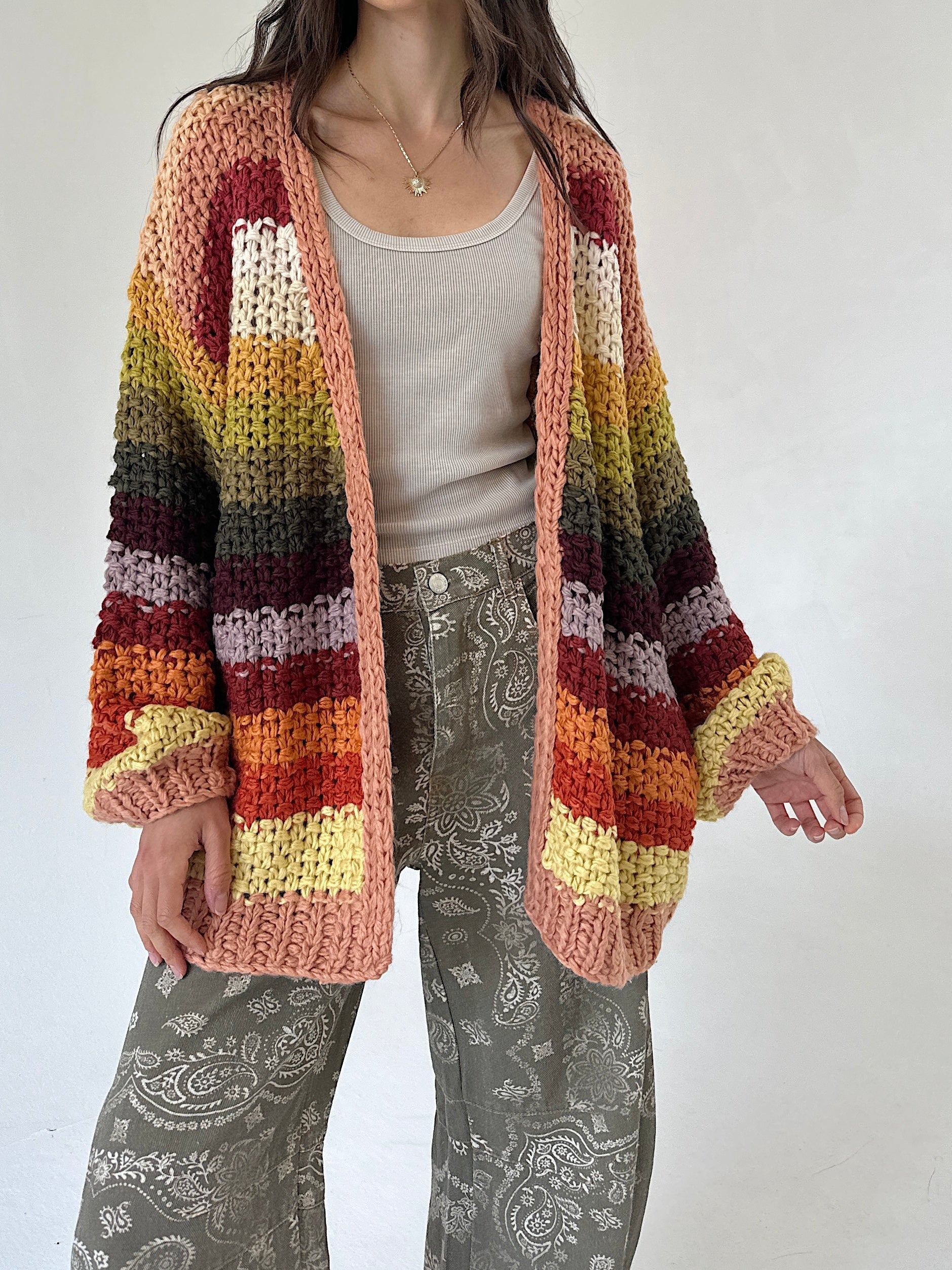 A person wearing a chunky knit open-front cardigan with horizontal multicolor stripes in peach, rust, orange, yellow, olive, and dark brown, layered over a light gray top and gray paisley pants against a plain light background.