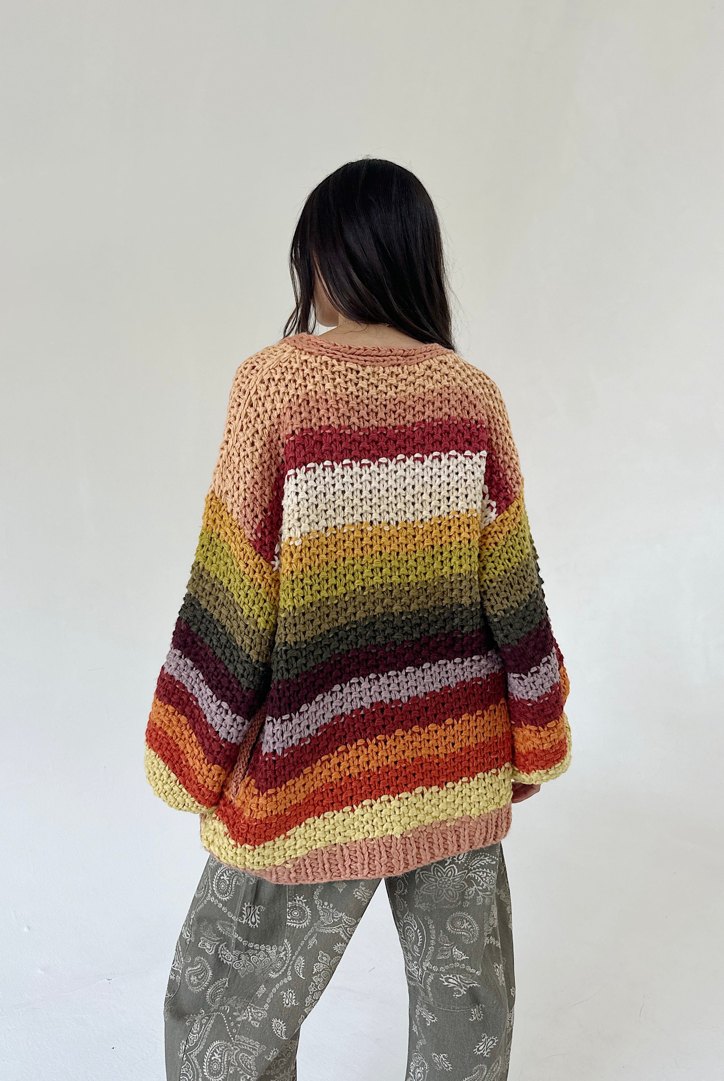 A person viewed from behind wearing a chunky knit striped sweater with horizontal bands of cream, yellow, green, brown, red, and orange, paired with olive patterned barrel pants, against a plain off-white wall.