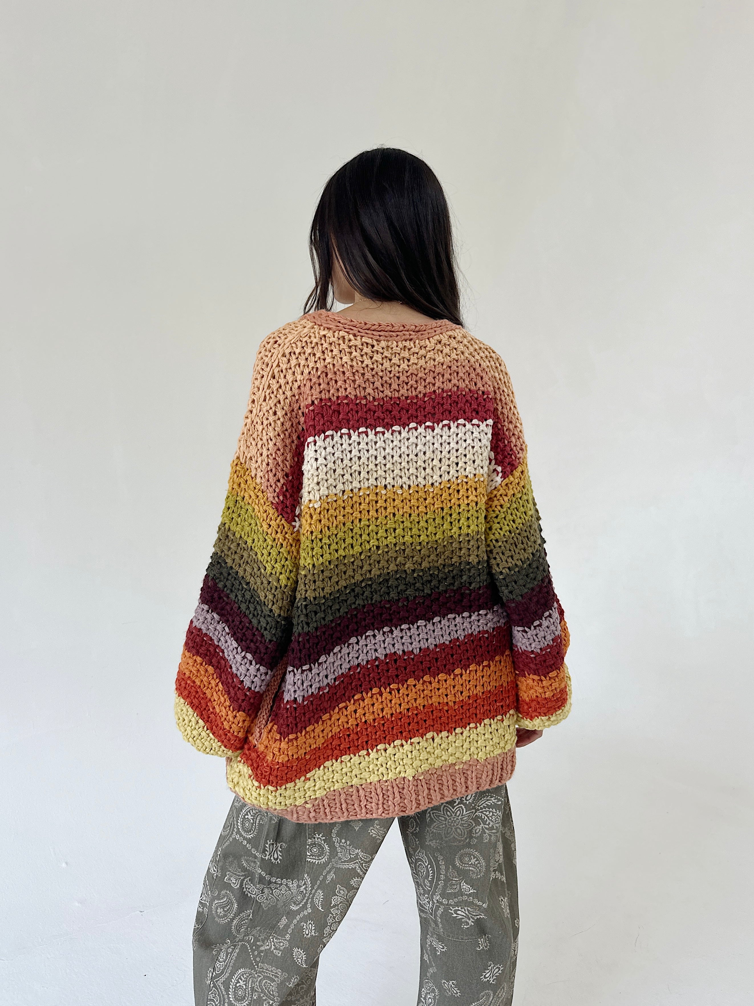 A person viewed from behind wearing a chunky knit striped sweater with horizontal bands of cream, yellow, green, brown, red, and orange, paired with olive patterned barrel pants, against a plain off-white wall.