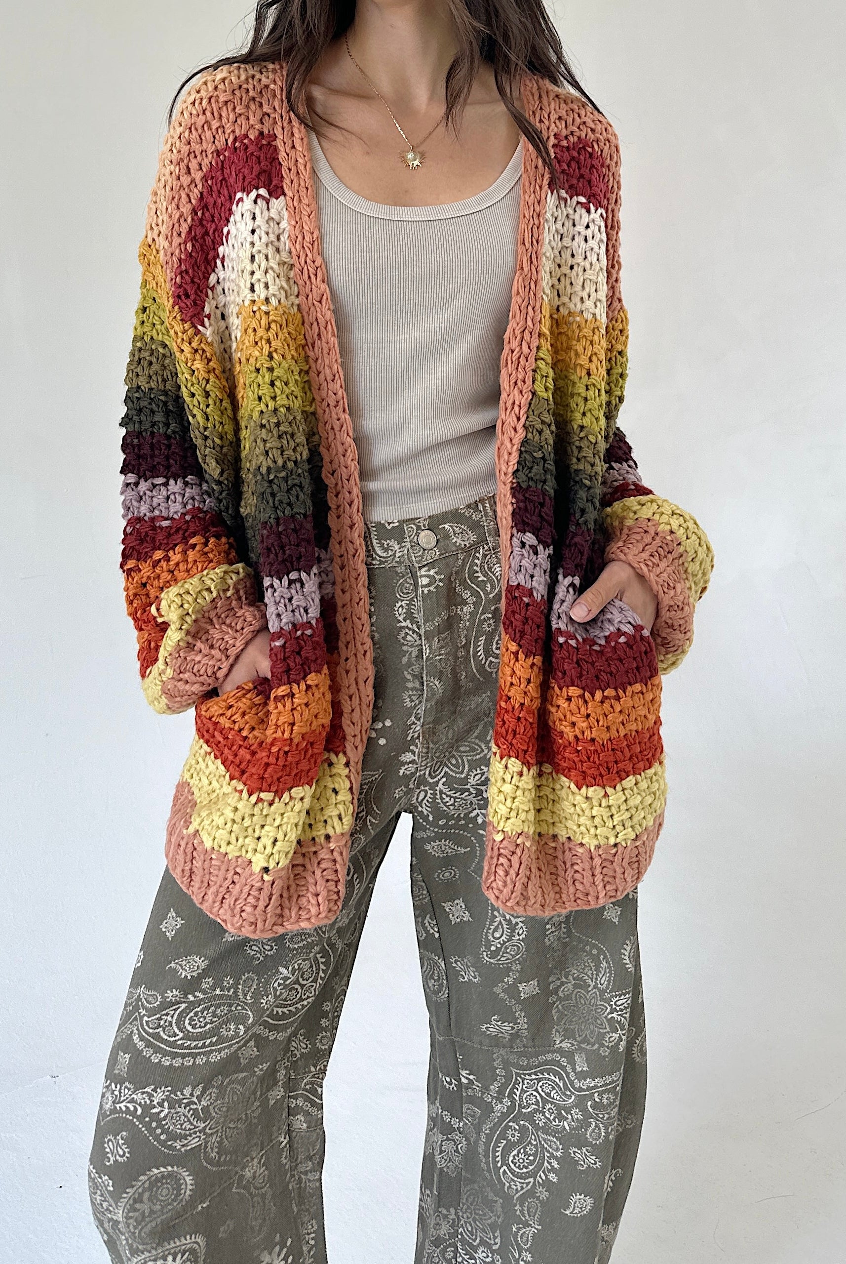 A person wearing a chunky striped crochet cardigan with horizontal bands of cream, yellow, orange, red, and brown, layered over a light gray tank top and gray paisley pants, against a plain off-white background.