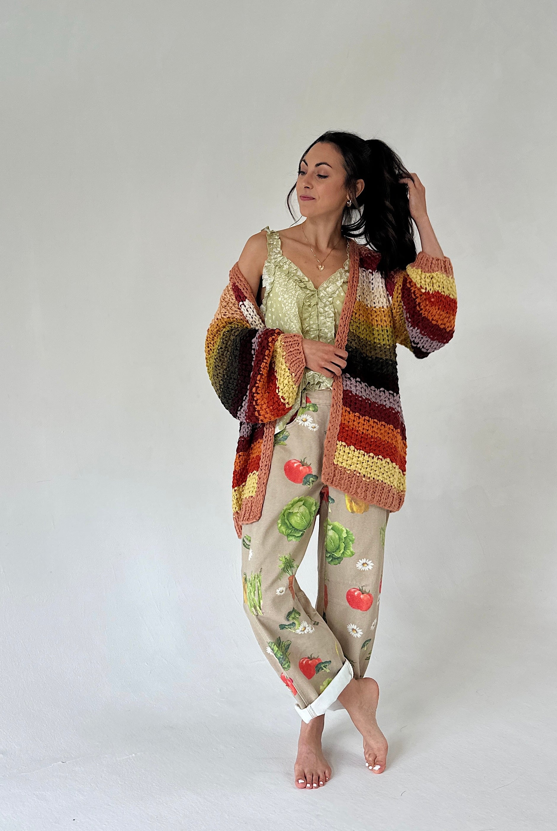 A woman stands barefoot against a plain light gray studio background wearing barrel pants with a vegetable print, a striped crochet cardigan in earthy tones, and a pale green lace-trim camisole.