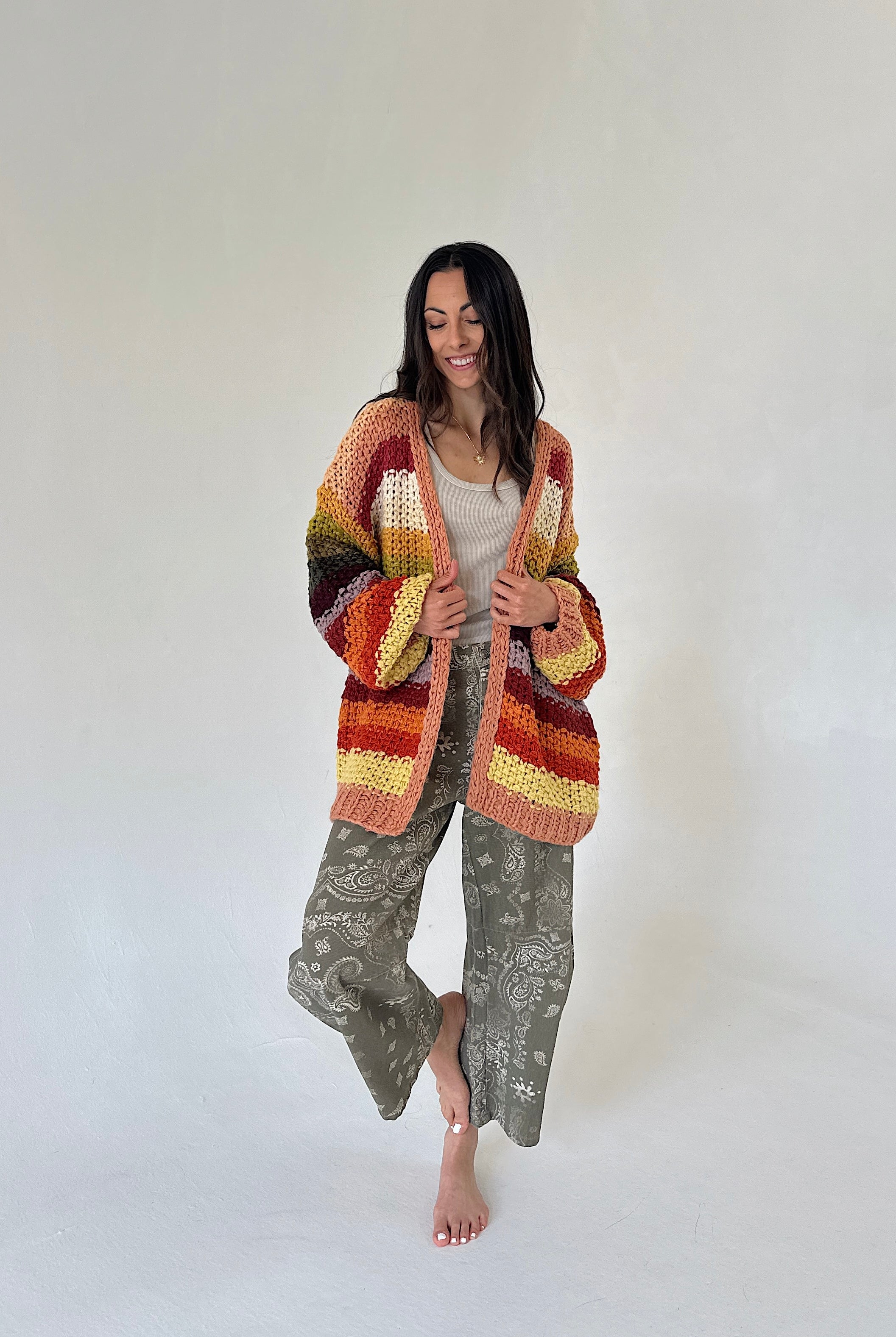 A woman stands barefoot against a plain light background wearing a chunky multicolor striped cardigan with rust, orange, yellow, and brown tones, a light beige top, and loose sage green bandana patterned barrel pants.