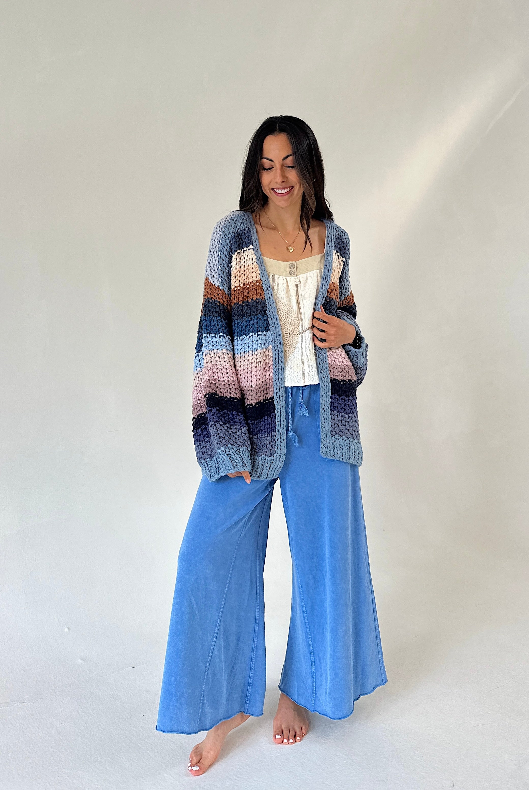 A woman stands barefoot against a plain light background wearing a chunky multicolor striped knit cardigan over a white top and wide-leg blue pants.
