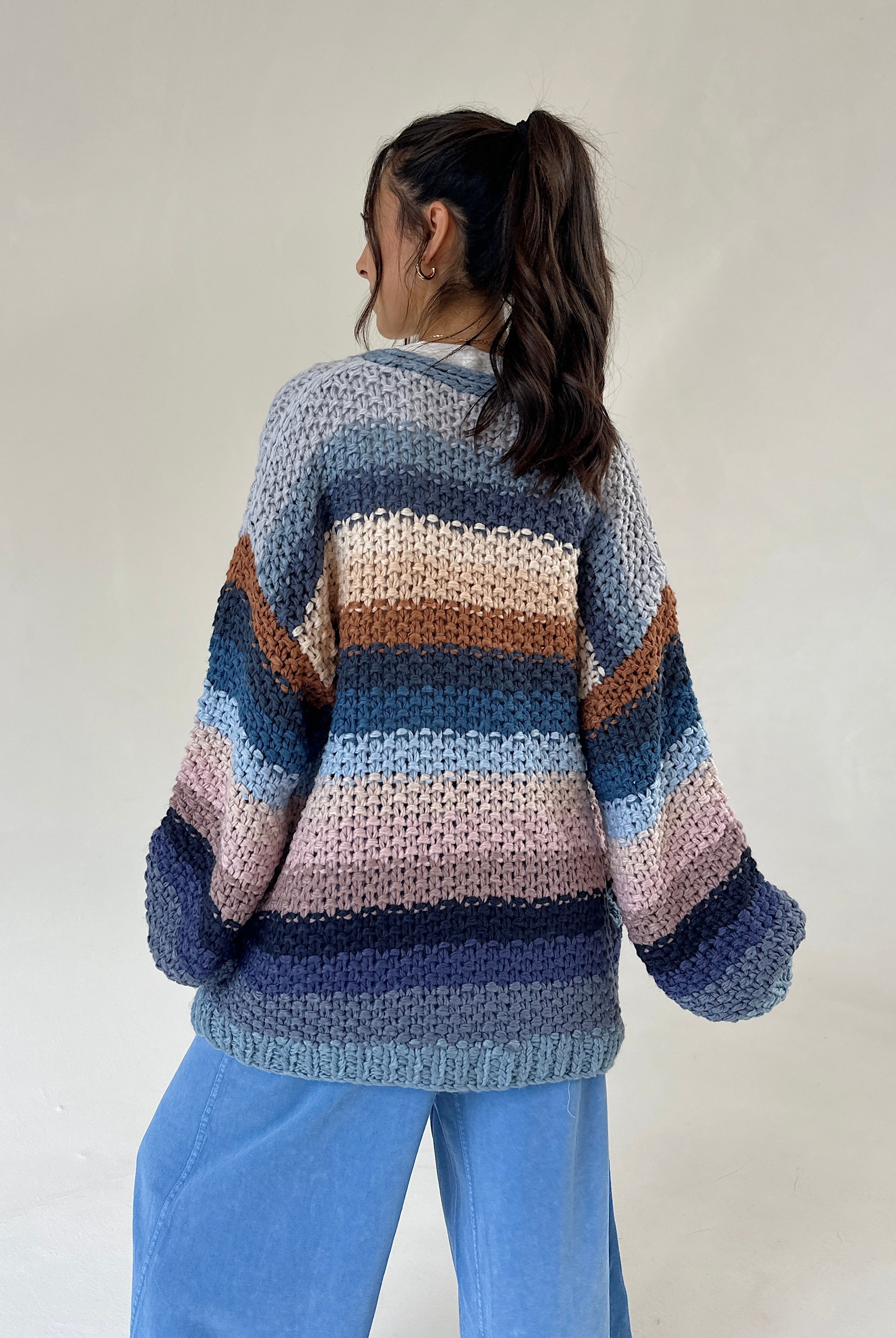 A person shown from the back wearing a chunky knit striped sweater with horizontal bands of blue, navy, beige, pink, and brown, paired with light blue pants, against a plain off-white background.