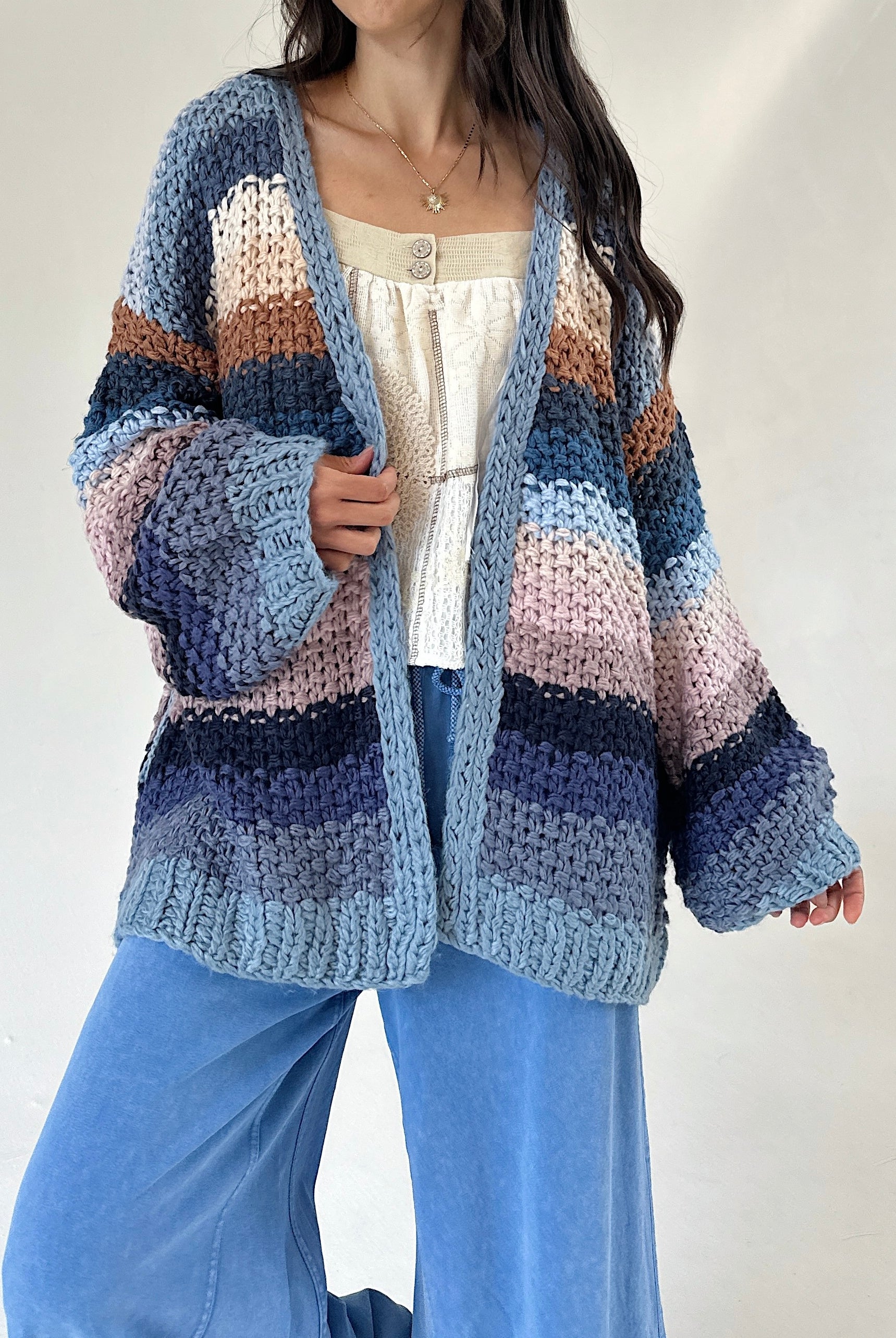 A woman wearing a chunky knit open-front cardigan with horizontal stripes in shades of blue, navy, beige, pink, and brown, styled over a cream top and blue wide-leg pants.