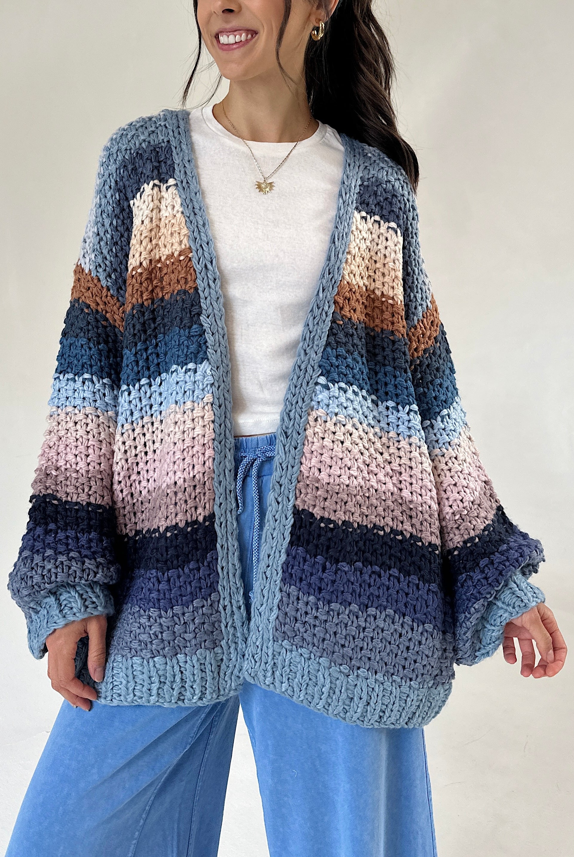 A woman wearing a chunky knit open-front cardigan with horizontal stripes in shades of blue, navy, pink, beige, and brown over a white top and blue pants, photographed against a plain light background.