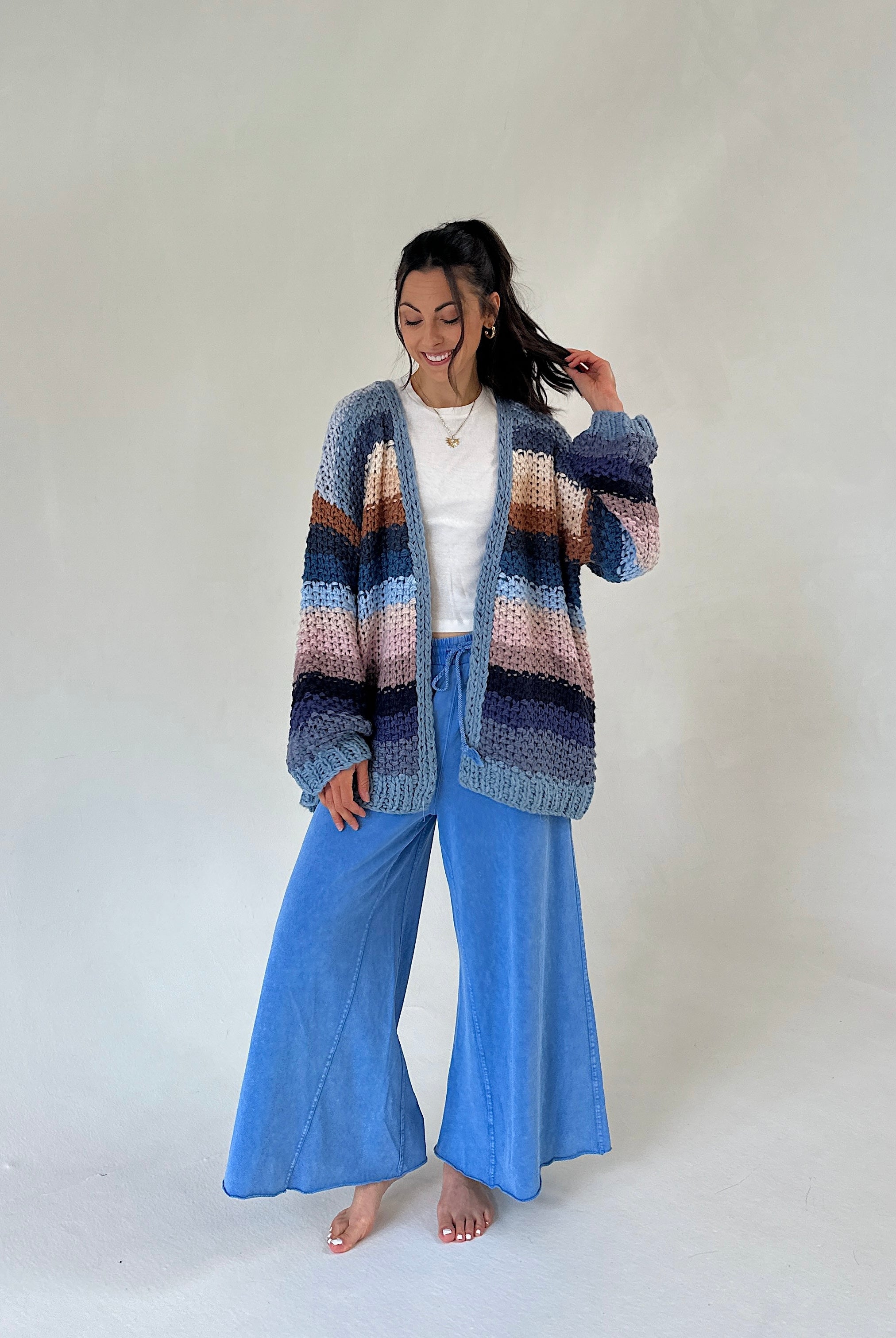 A woman stands barefoot against a plain light gray studio background, wearing a chunky knit cardigan with horizontal stripes in blue, purple, pink, and brown over a white top and wide-leg blue pants.