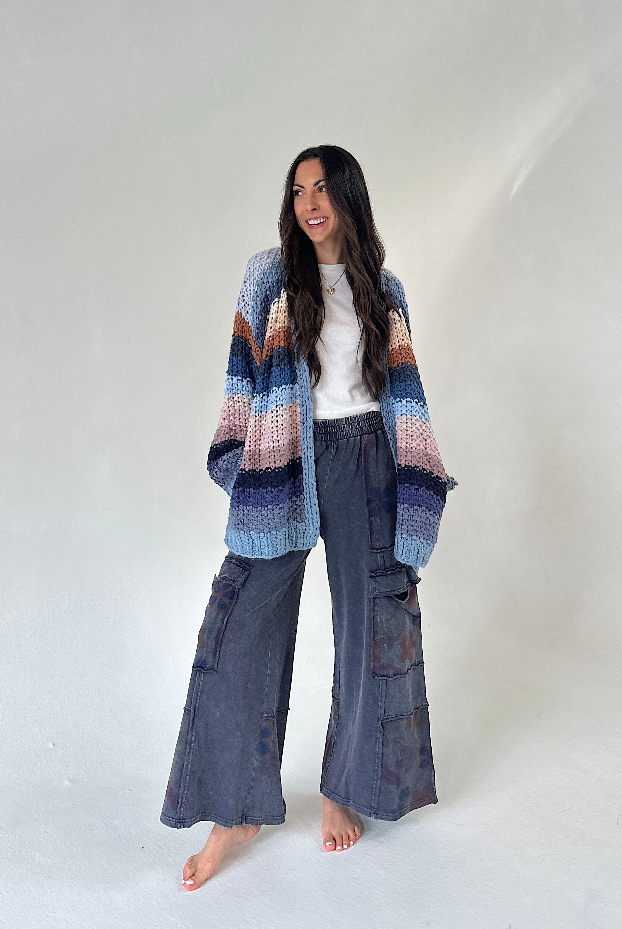 A woman stands barefoot against a plain light gray studio backdrop, wearing a loose multicolor striped knit cardigan over a white top and dark gray wide-leg cargo pants.