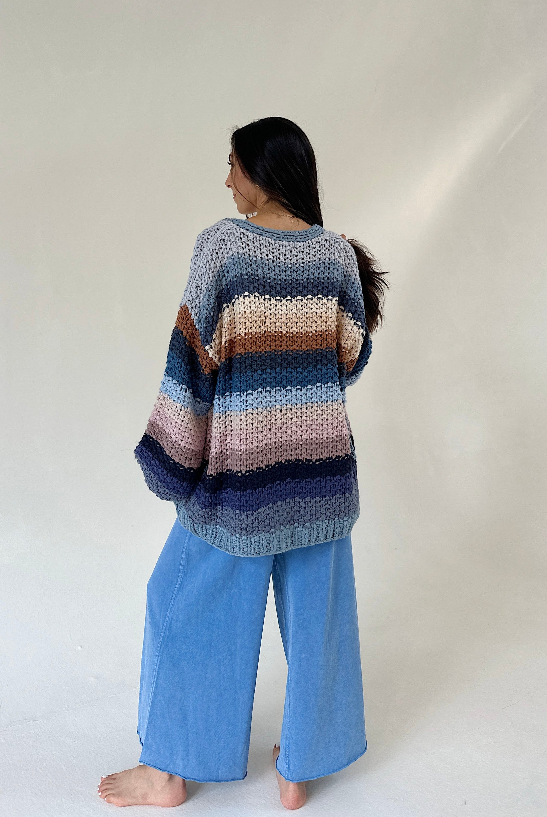 A woman stands barefoot against a plain light background, viewed from the back, wearing a loose striped knit sweater with blue wide-leg pants.