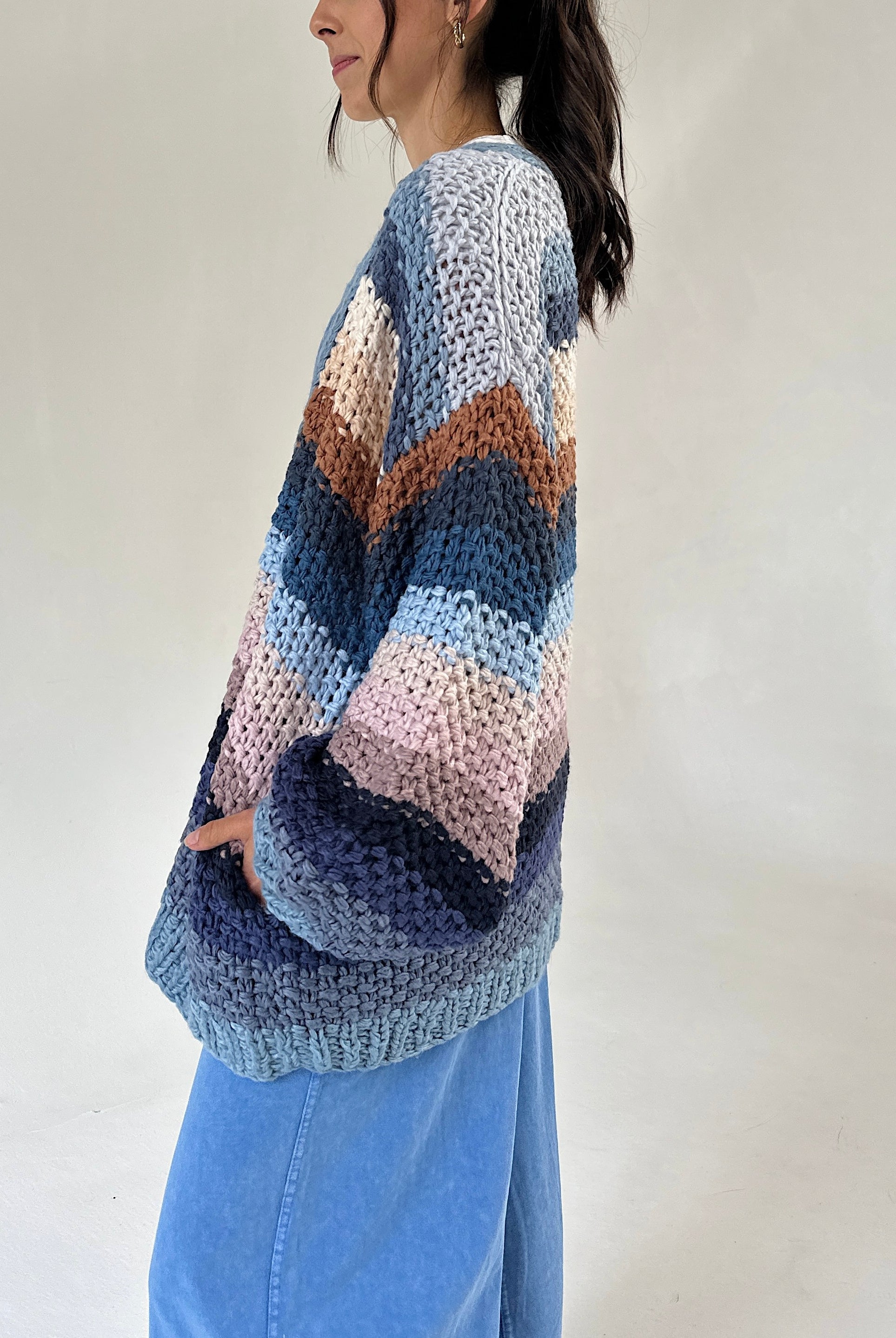 A woman wearing a chunky cardigan with horizontal stripes in shades of blue, navy, pink, and brown, paired with light blue jeans against a plain off-white background.
