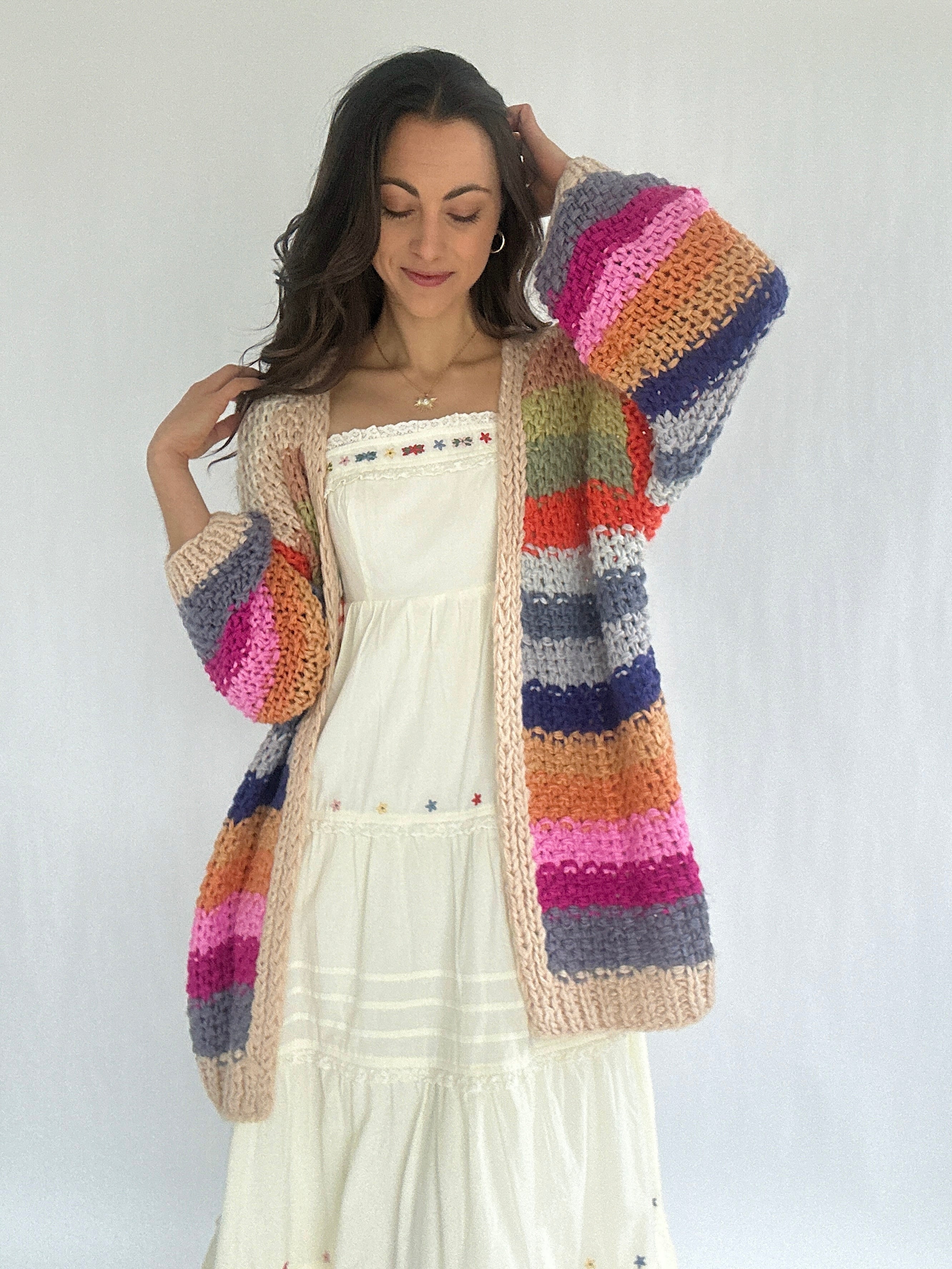 Woman wearing a colorful striped crochet cardigan over a white dress against a plain background