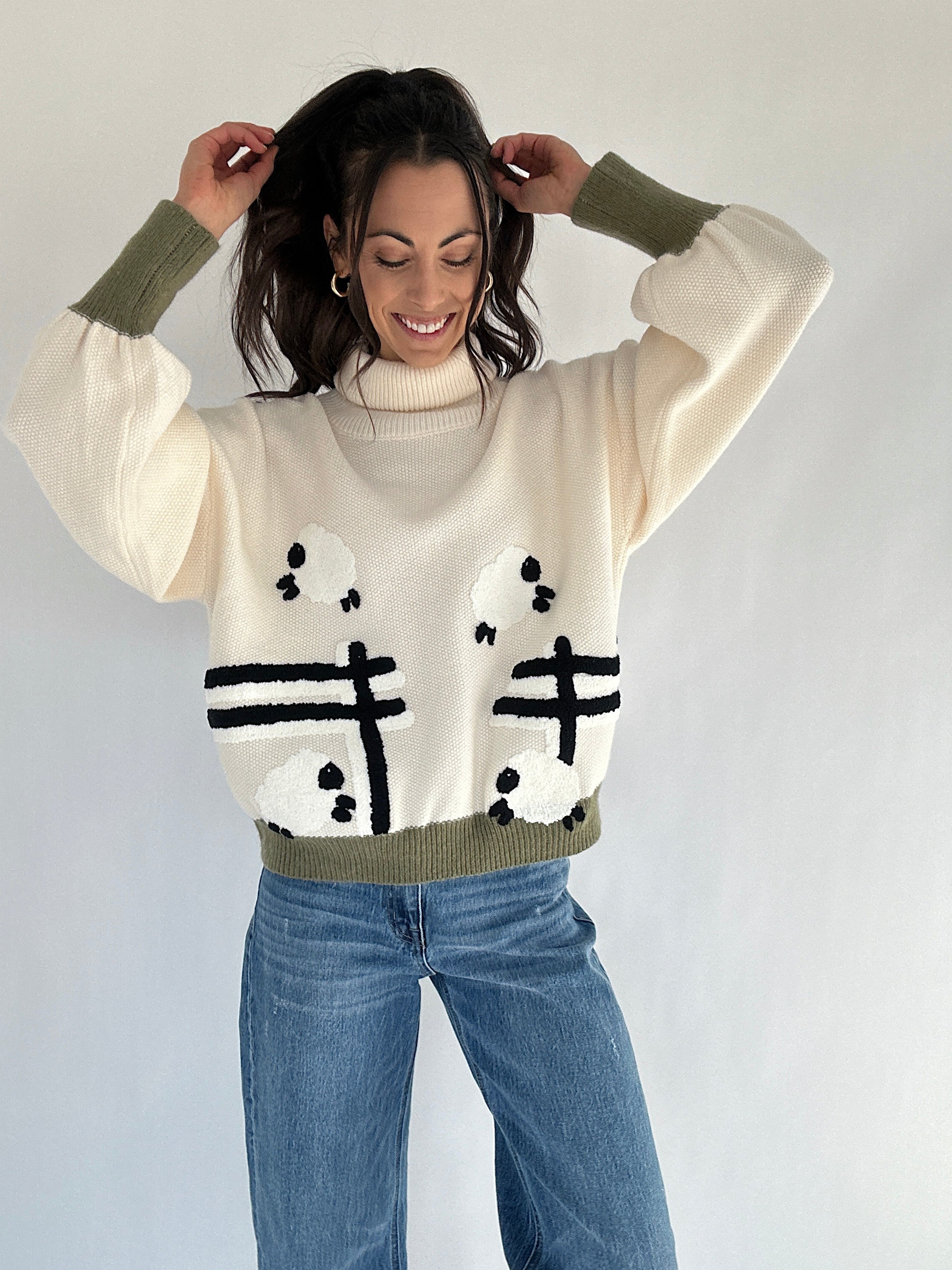 Woman wearing a cream sweater with sheep jumping over a fence and green trim, standing against a plain background