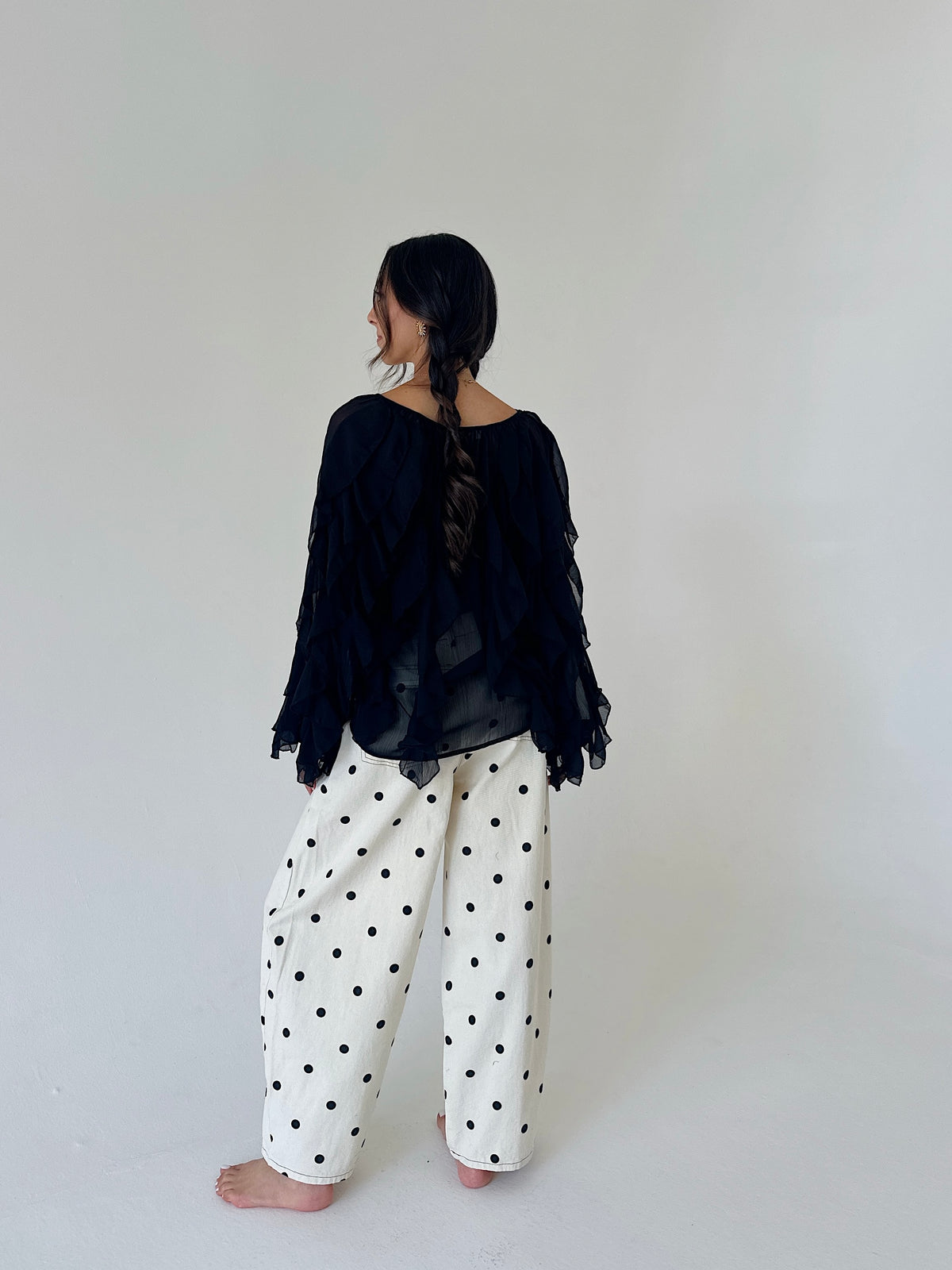 A woman stands barefoot against a plain light gray studio background, viewed from behind. She is wearing a loose black sheer long-sleeve top with ruffled edges and wide-leg white pants covered in black polka dots.