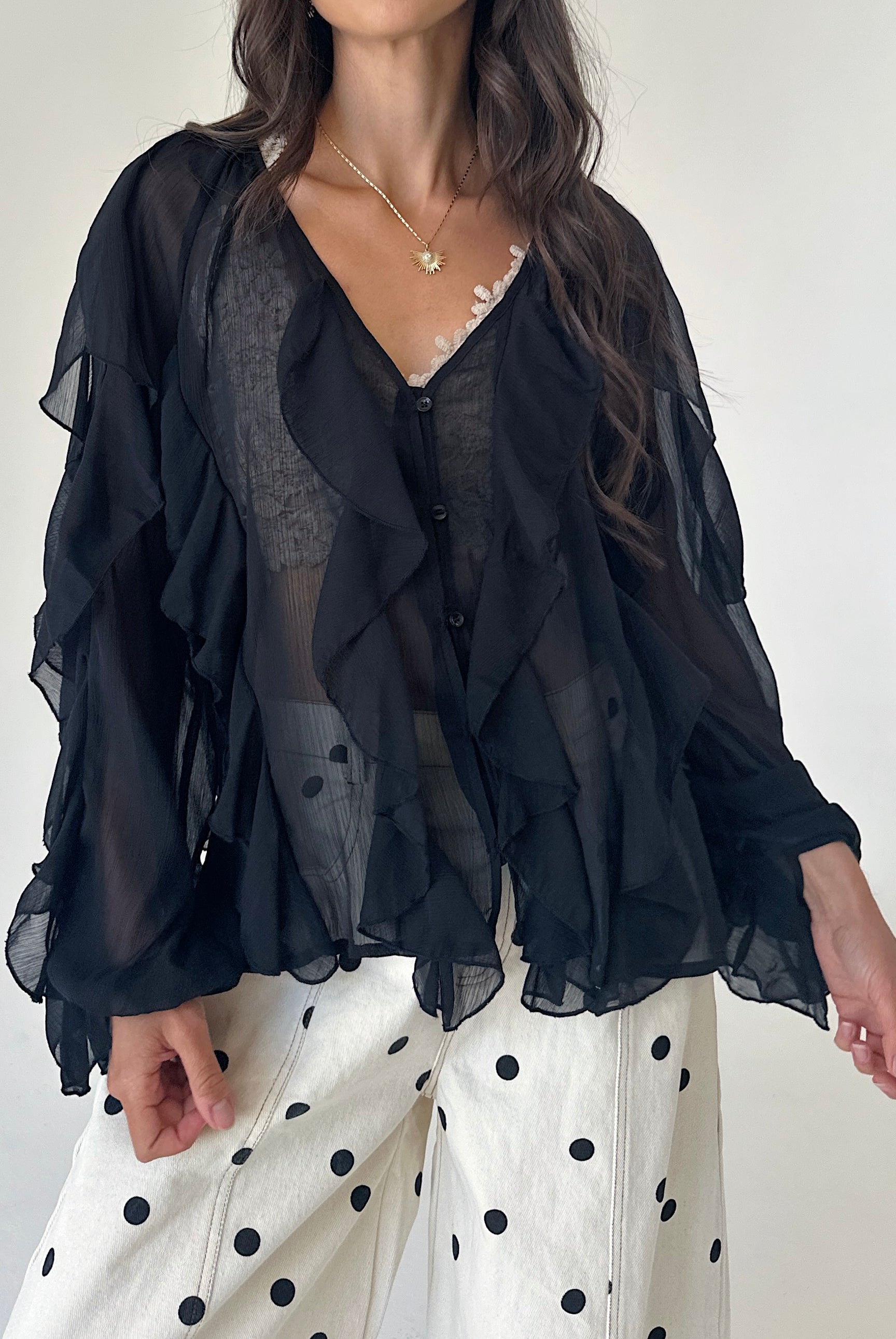 A woman wearing a sheer black ruffled blouse over a gray lace top, paired with white pants featuring black polka dots, against a plain off-white background.