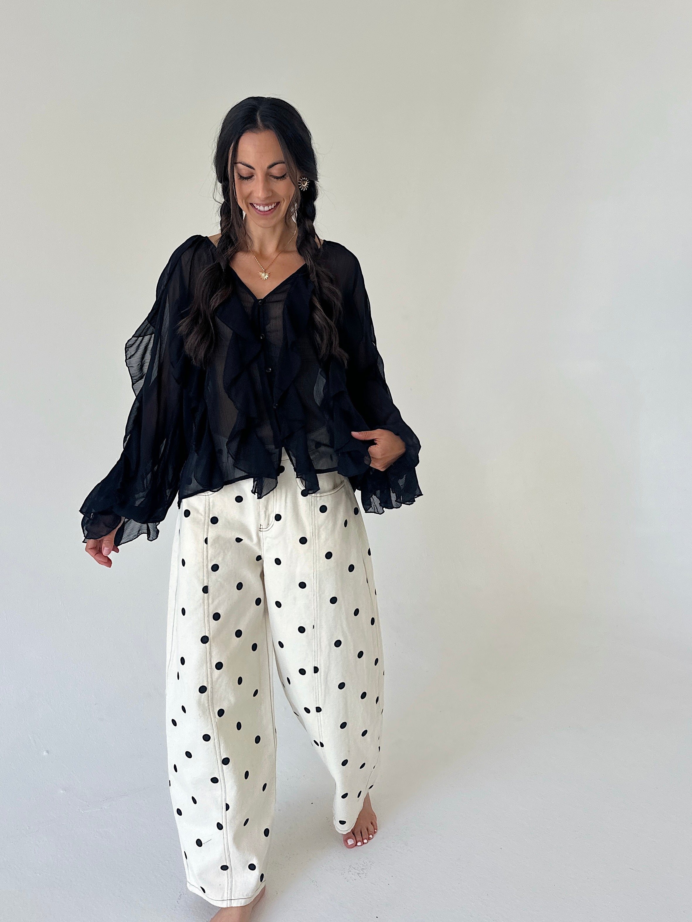 A woman modeling a black sheer long-sleeve blouse with ruffles and wide-leg white pants with black polka dots, standing against a plain off-white studio background.