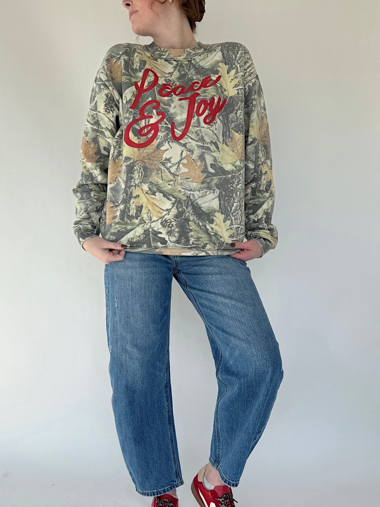 Person wearing a camouflage sweatshirt with 'Peace & Joy' text and blue jeans on a white background
