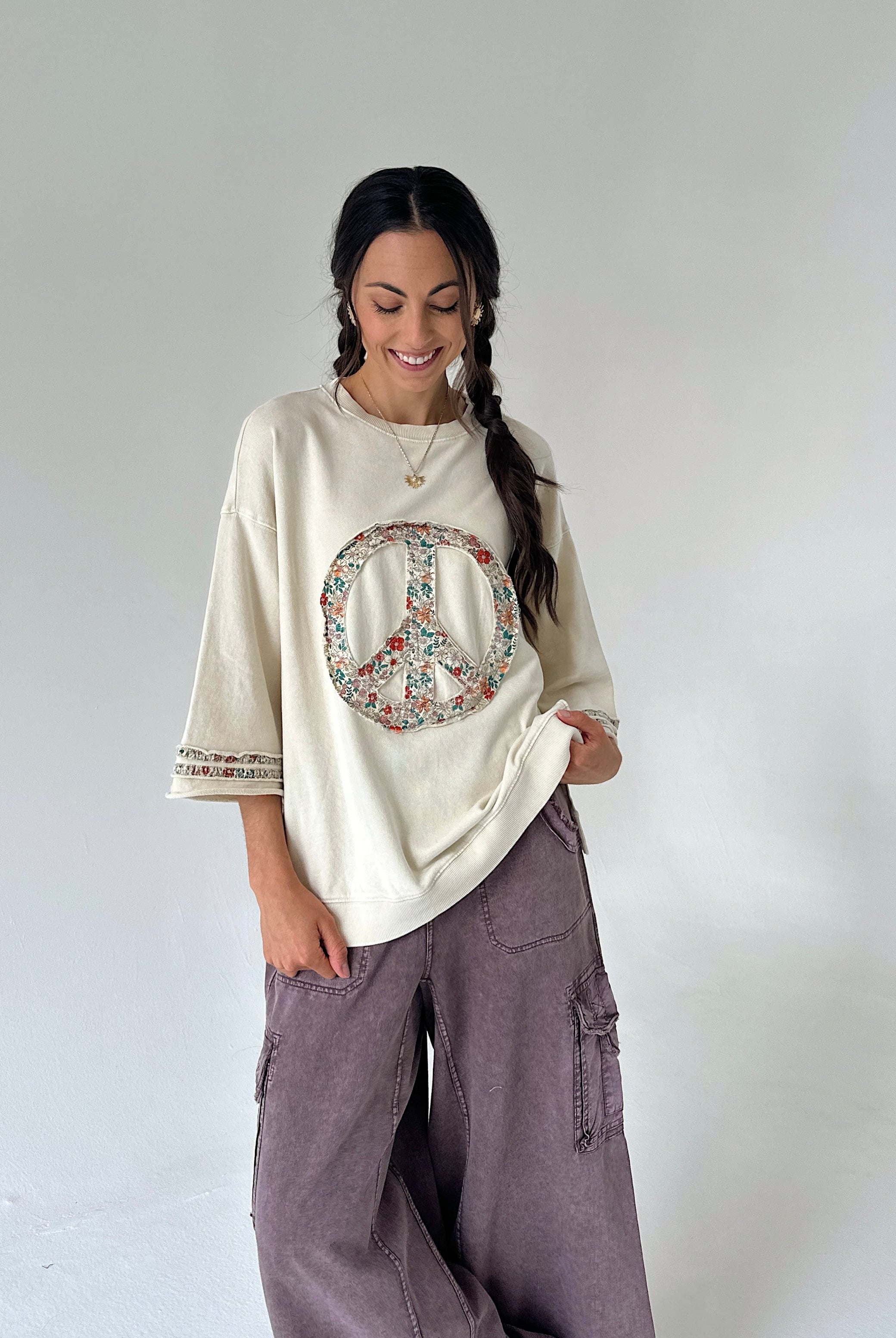 A woman modeling a cream-colored oversized t-shirt with a colorful peace sign graphic on the front, paired with wide-leg mauve cargo pants, against a plain light gray studio background.