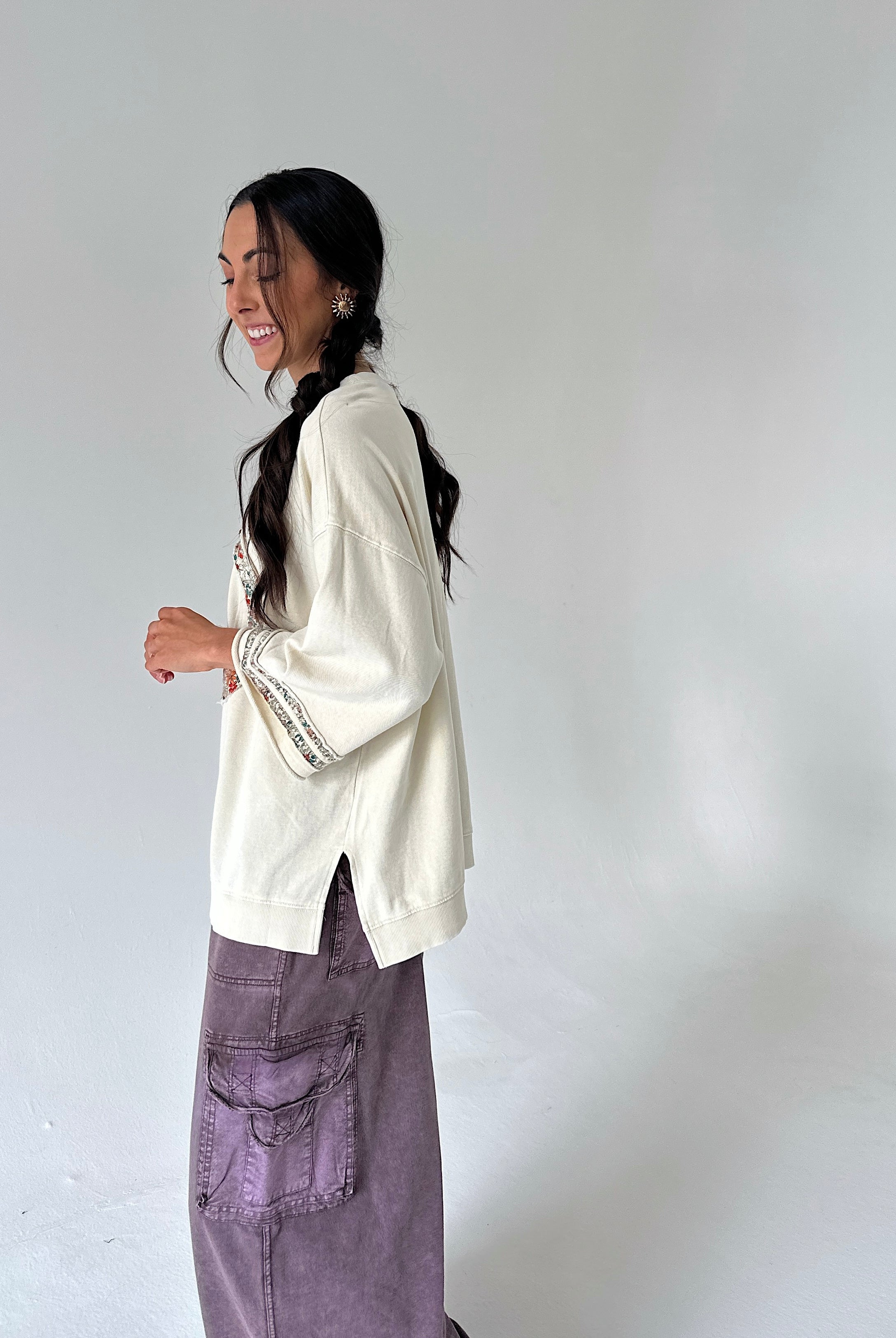 A woman wearing a cream-colored knit sweater with embroidered details on the sleeve and a purple cargo skirt, standing against a plain light gray studio background.