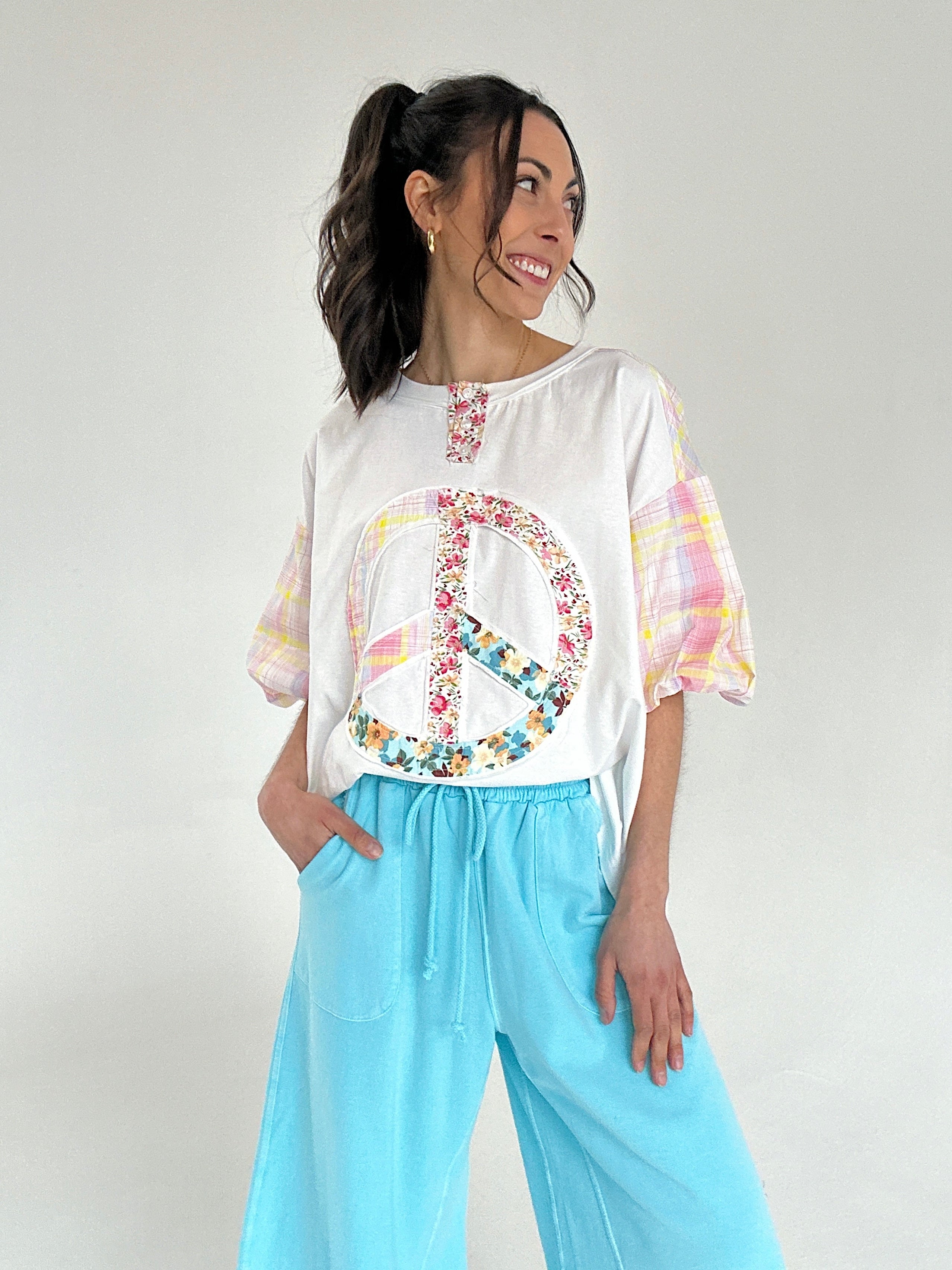Woman wearing a white blouse with colorful patterns and light blue pants on a plain background