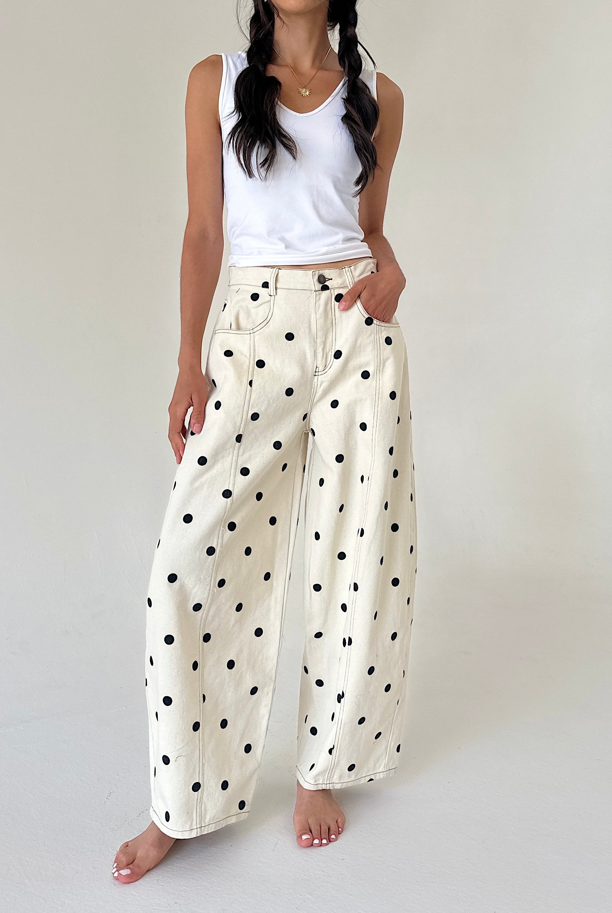 A model wearing cream wide-leg pants with a black polka dot pattern, styled with a white ribbed tank top, standing against a neutral off-white studio background.