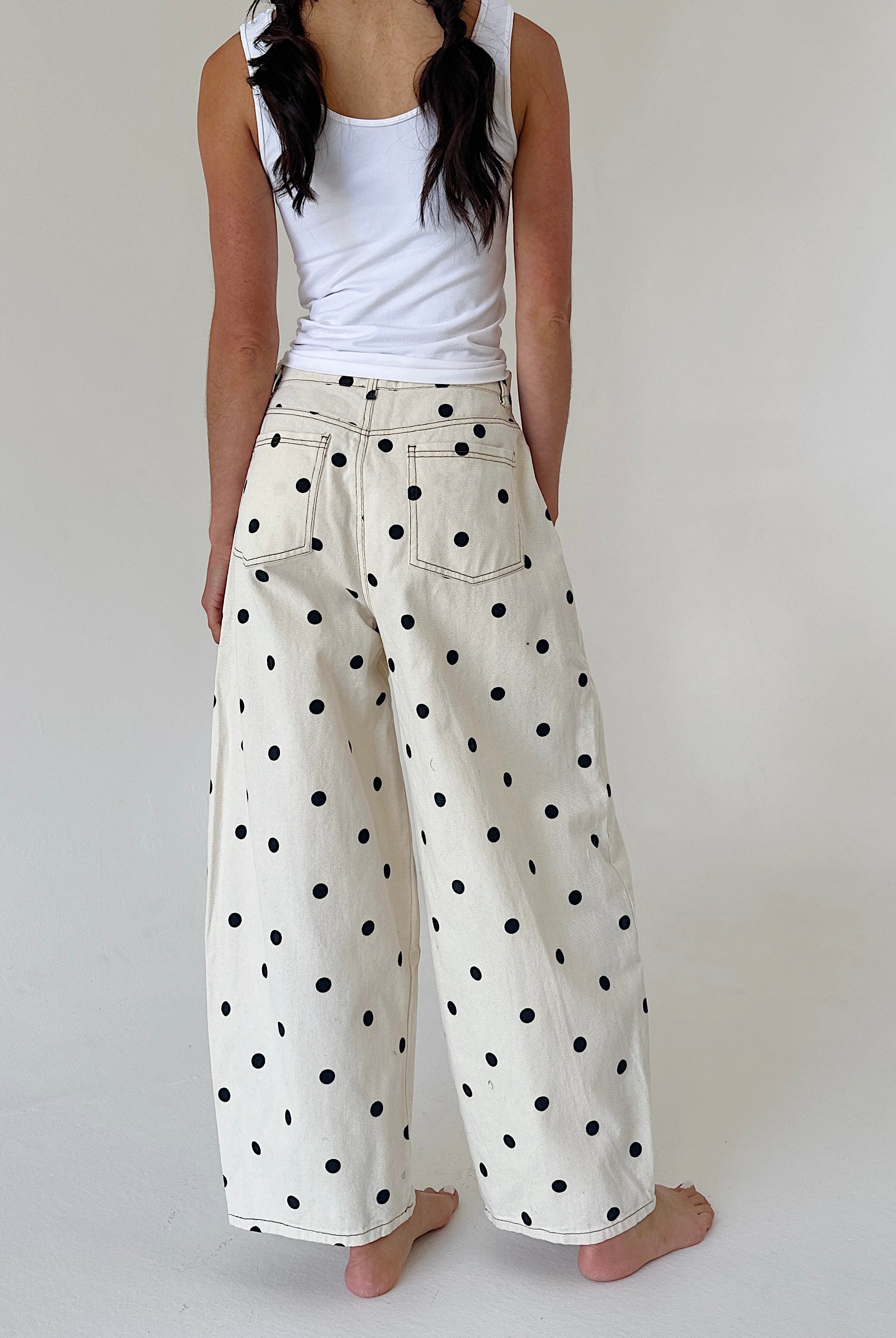 A person shown from the back wearing wide-leg cream pants with black polka dots and a white tank top, standing barefoot against a plain light background.