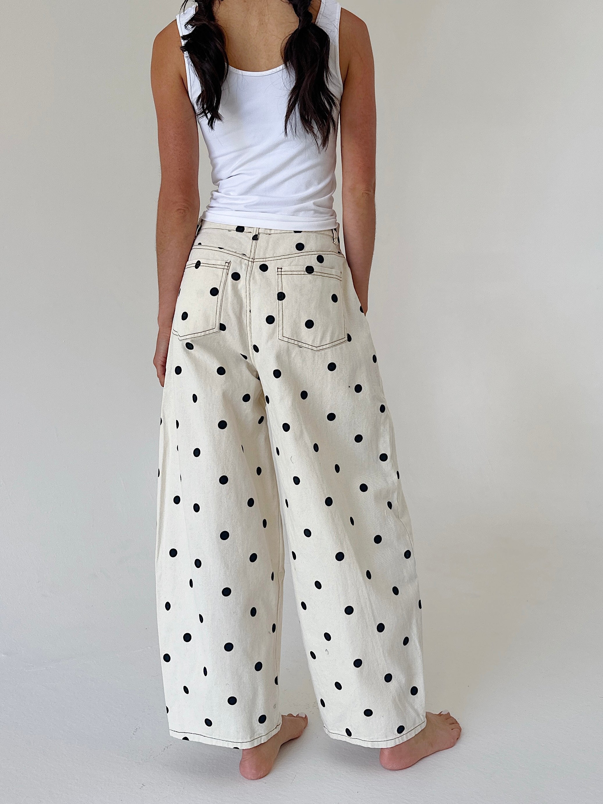 A person shown from the back wearing wide-leg cream pants with black polka dots and a white tank top, standing barefoot against a plain light background.