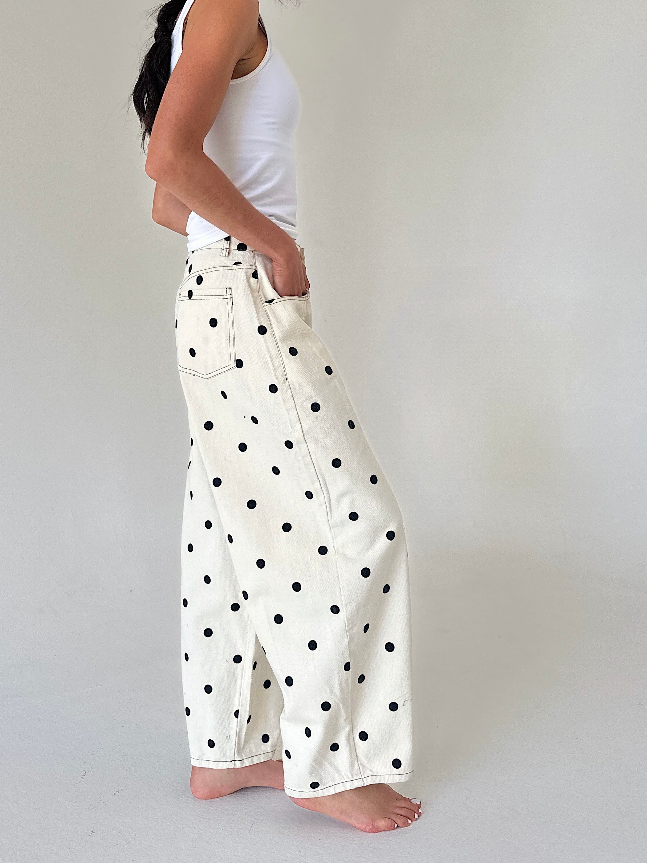 A person wearing off-white wide-leg pants with black polka dots, paired with a white tank top, standing barefoot against a plain light gray studio background.