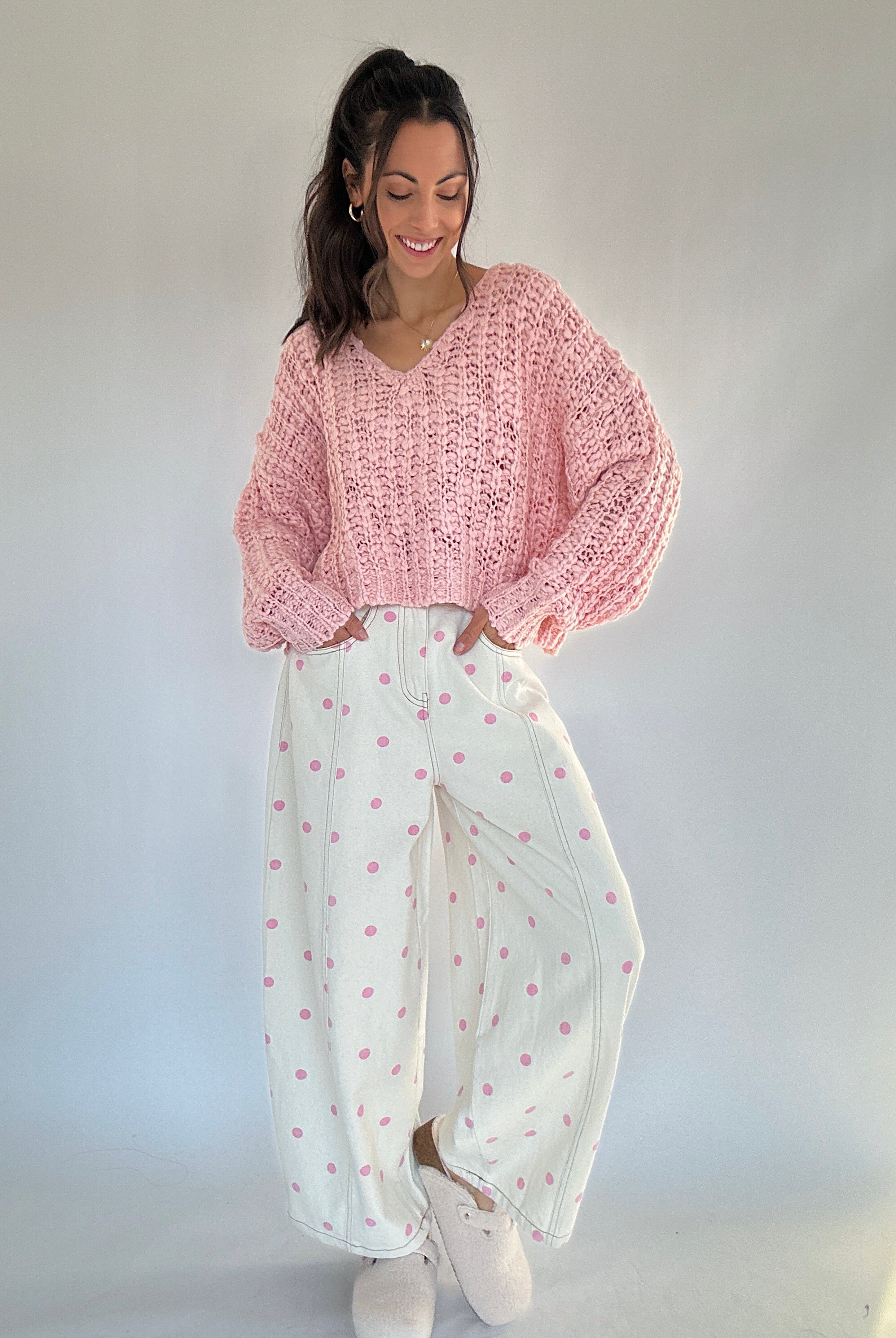 Woman wearing a pink knitted sweater and white pants with pink polka dot patterns against a plain background