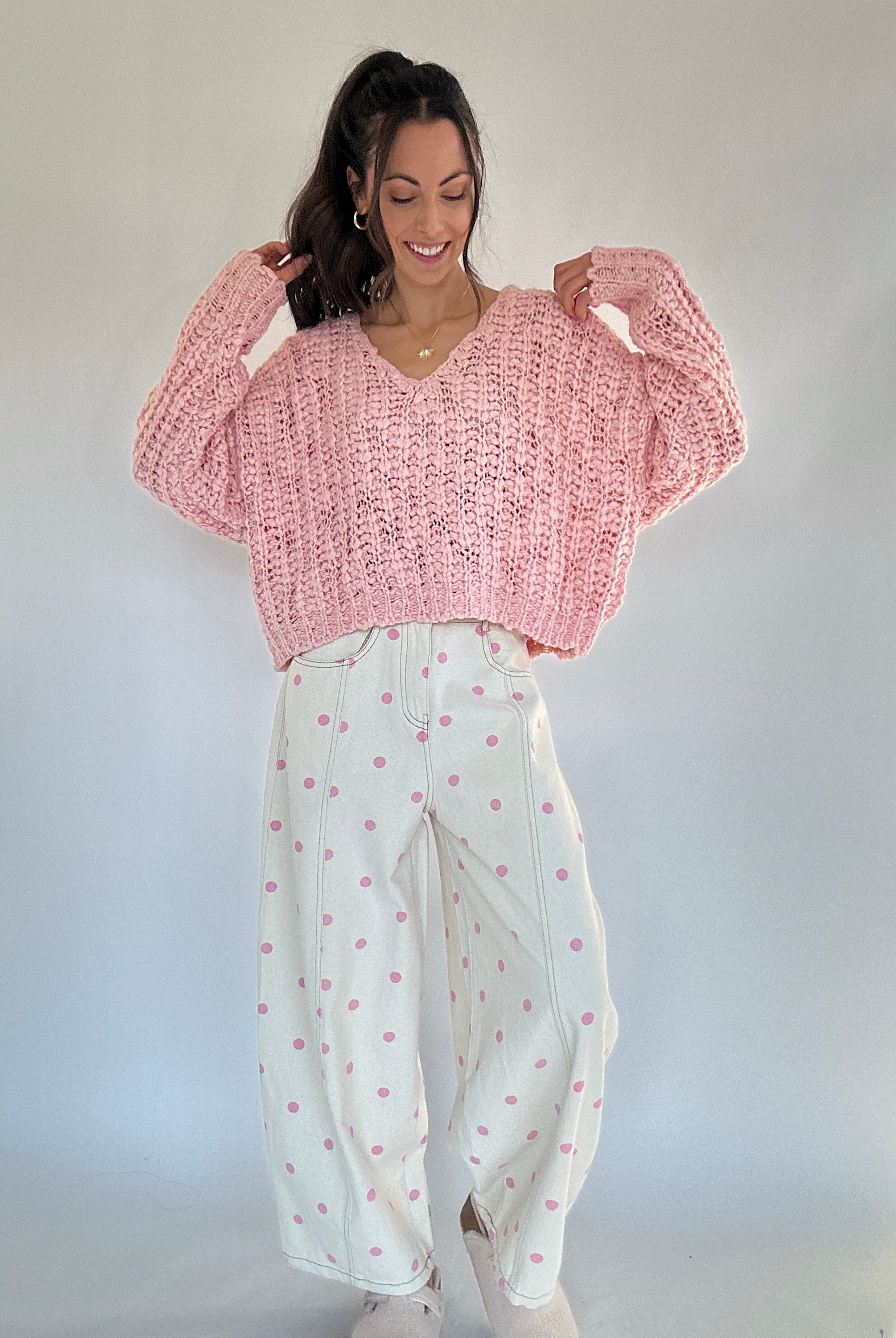 Woman wearing a pink knitted sweater and white pants with pink polka dot patterns against a plain background