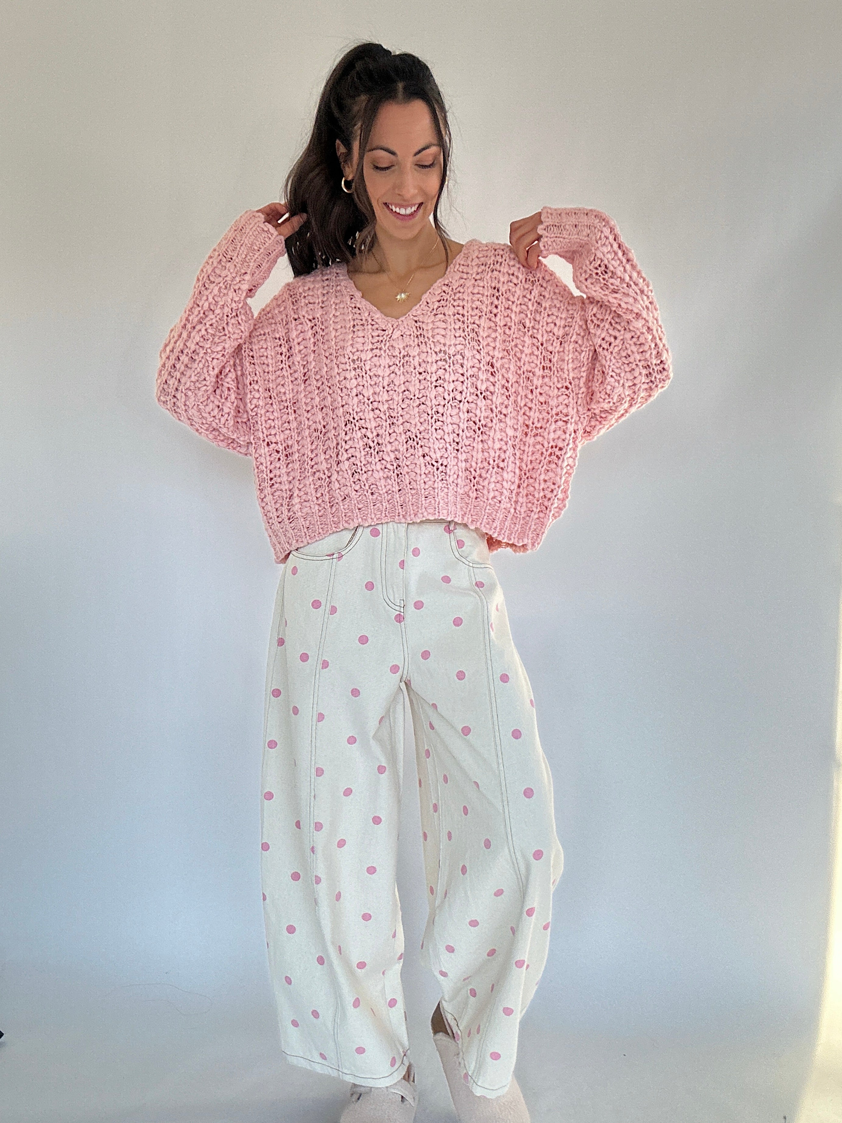 Woman wearing a pink knitted sweater and white pants with pink polka dot patterns against a plain background