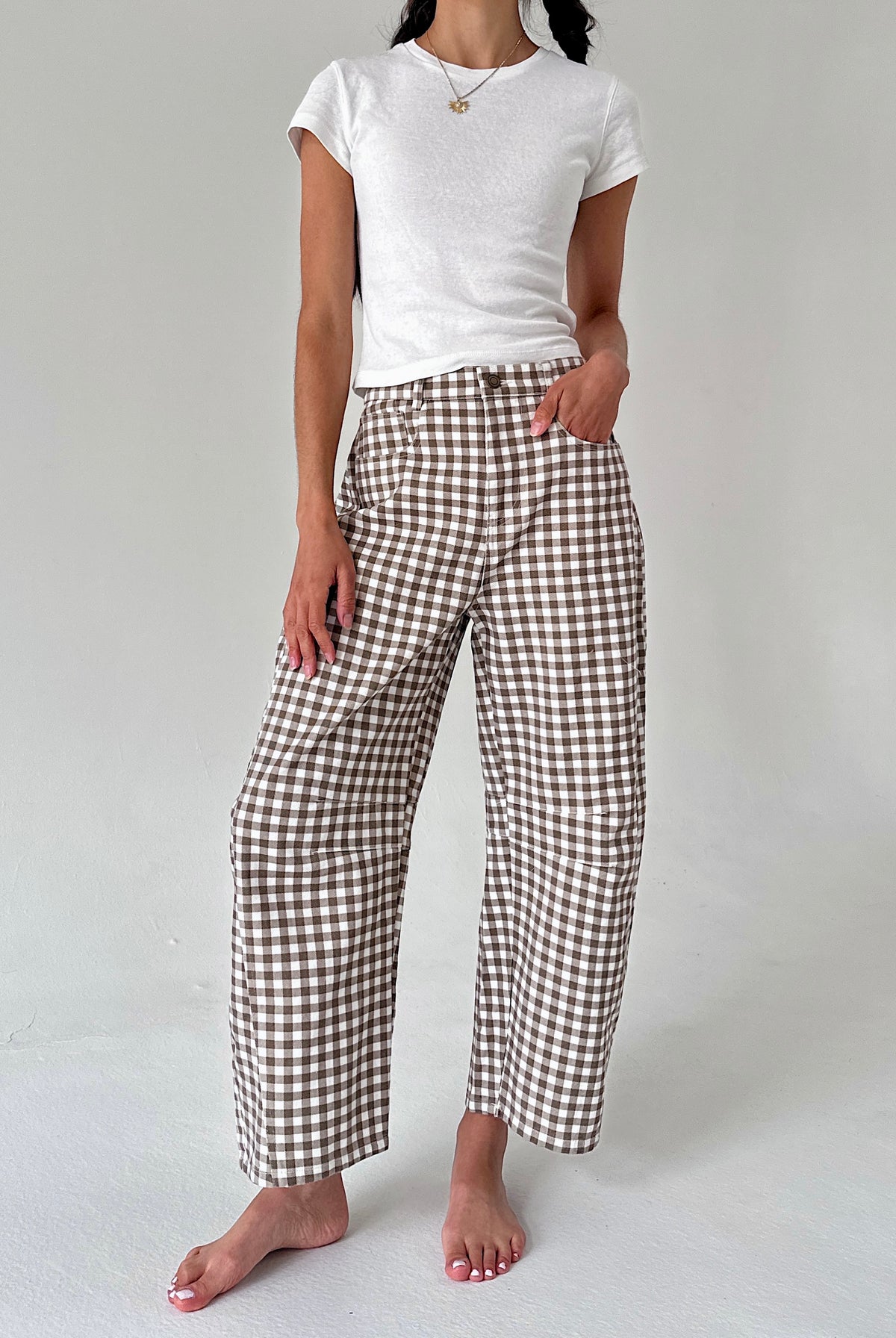 A model wearing brown and white gingham wide-leg cropped pants with a white t-shirt, standing barefoot against a light gray studio background.