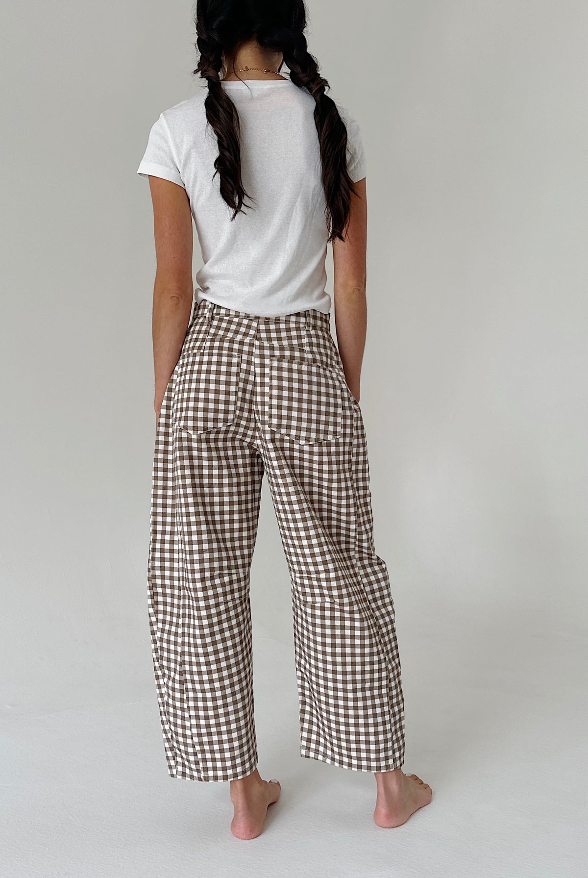 A person shown from the back wearing brown and white gingham wide-leg pants with a white t-shirt, standing barefoot against a light gray studio background.