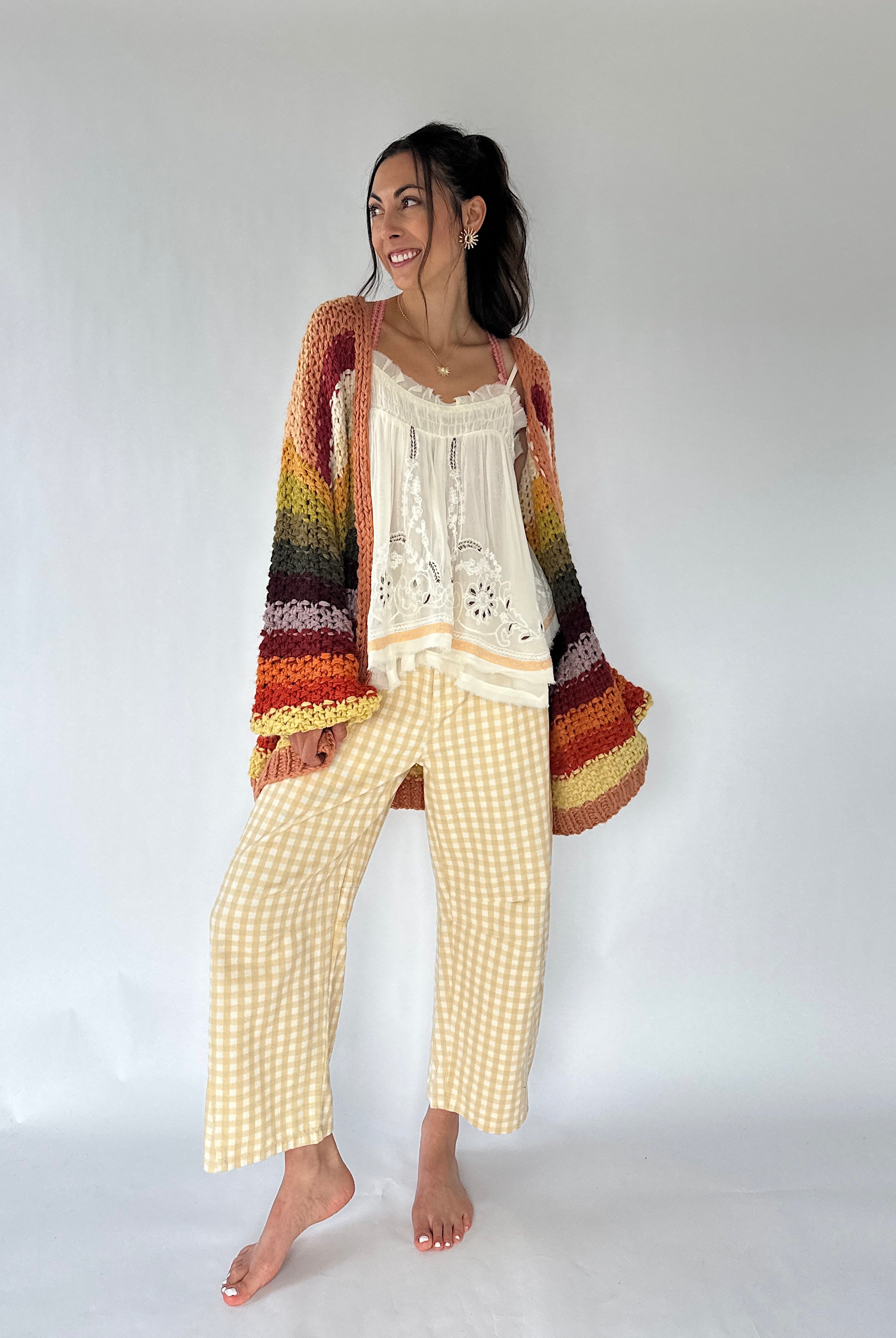 A woman modeling a cozy bohemian-style outfit consisting of a multicolor striped crochet cardigan and a cream crochet top paired with yellow-and-white gingham cropped pants, against a plain white studio background.