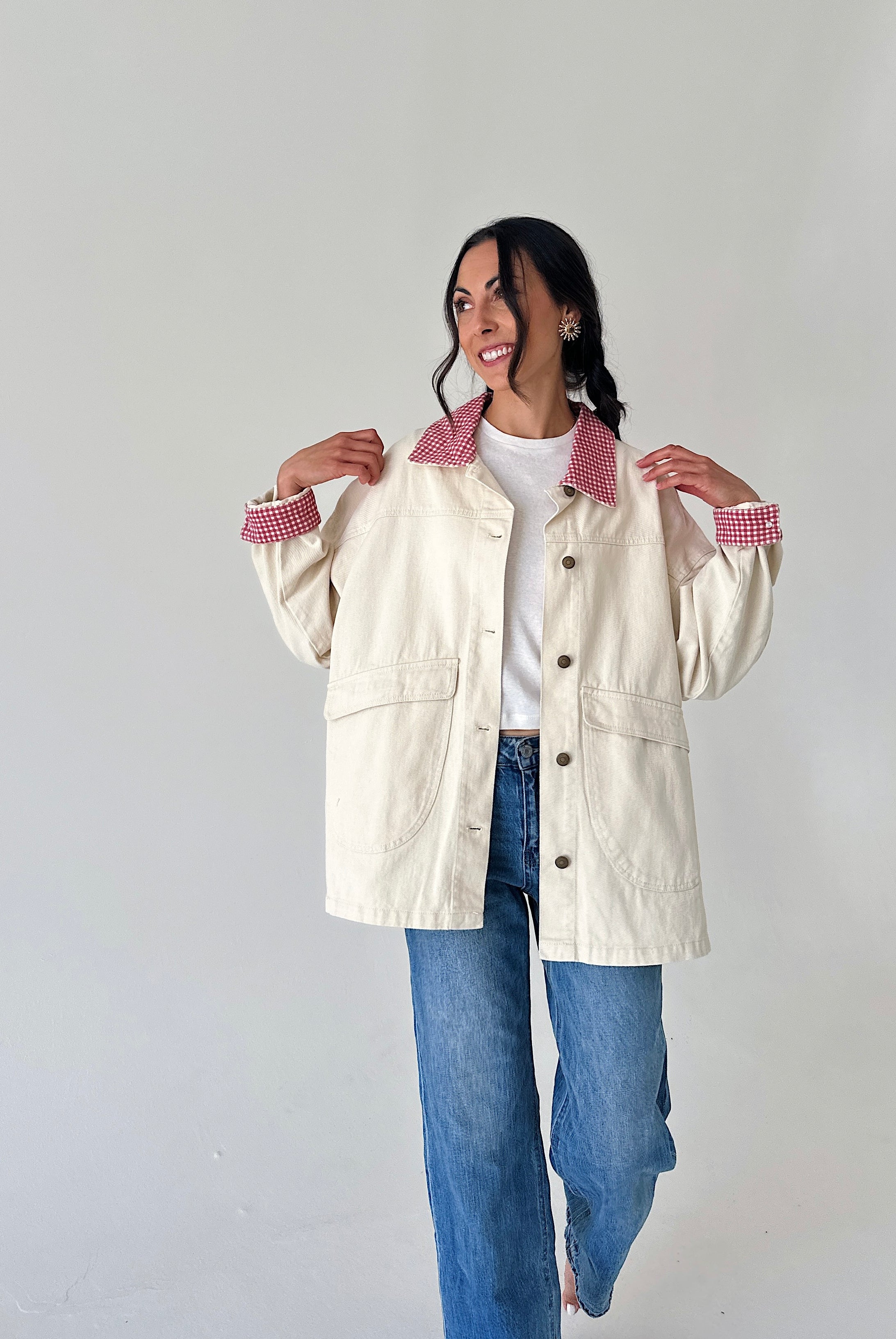 A woman modeling a cream-colored utility jacket with red-and-white gingham patterned collars and cuffs, worn over a white t-shirt and blue jeans, against a plain light gray background.