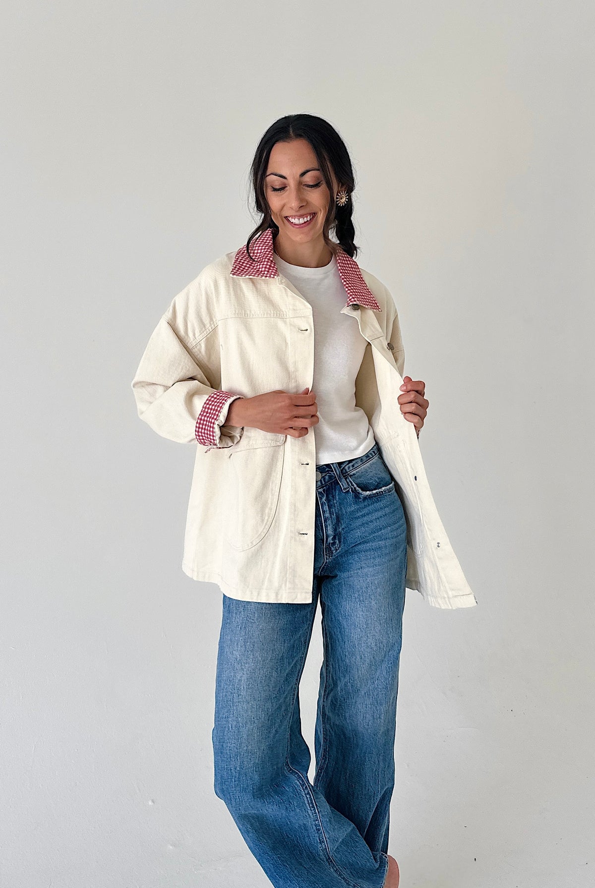 A woman modeling a cream-colored utility jacket with a pink and white gingham patterned collar and cuffs, worn over a white t-shirt and blue jeans, against a plain light gray background.