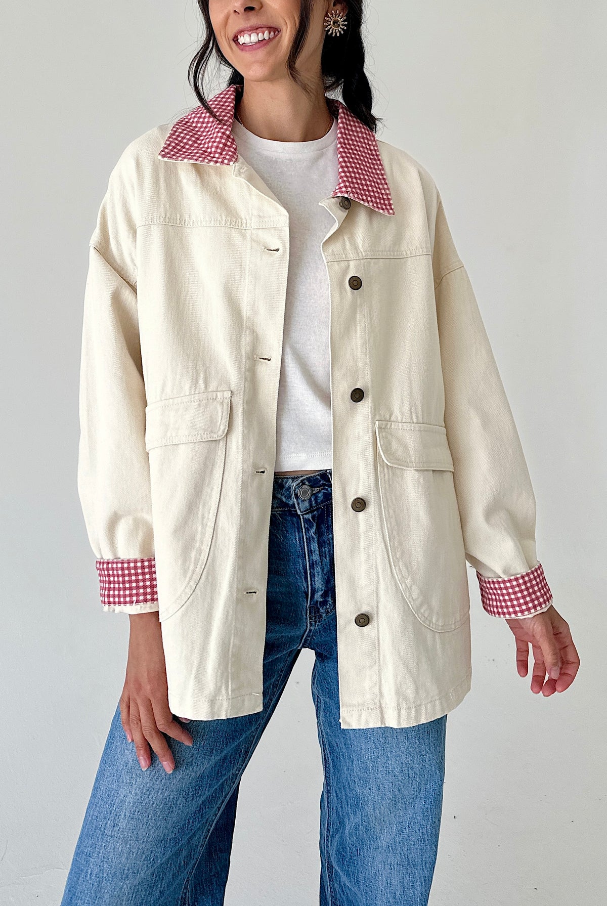 A woman wearing a cream-colored utility jacket with red and white gingham patterned collar and cuffs, paired with a white t-shirt and blue jeans.