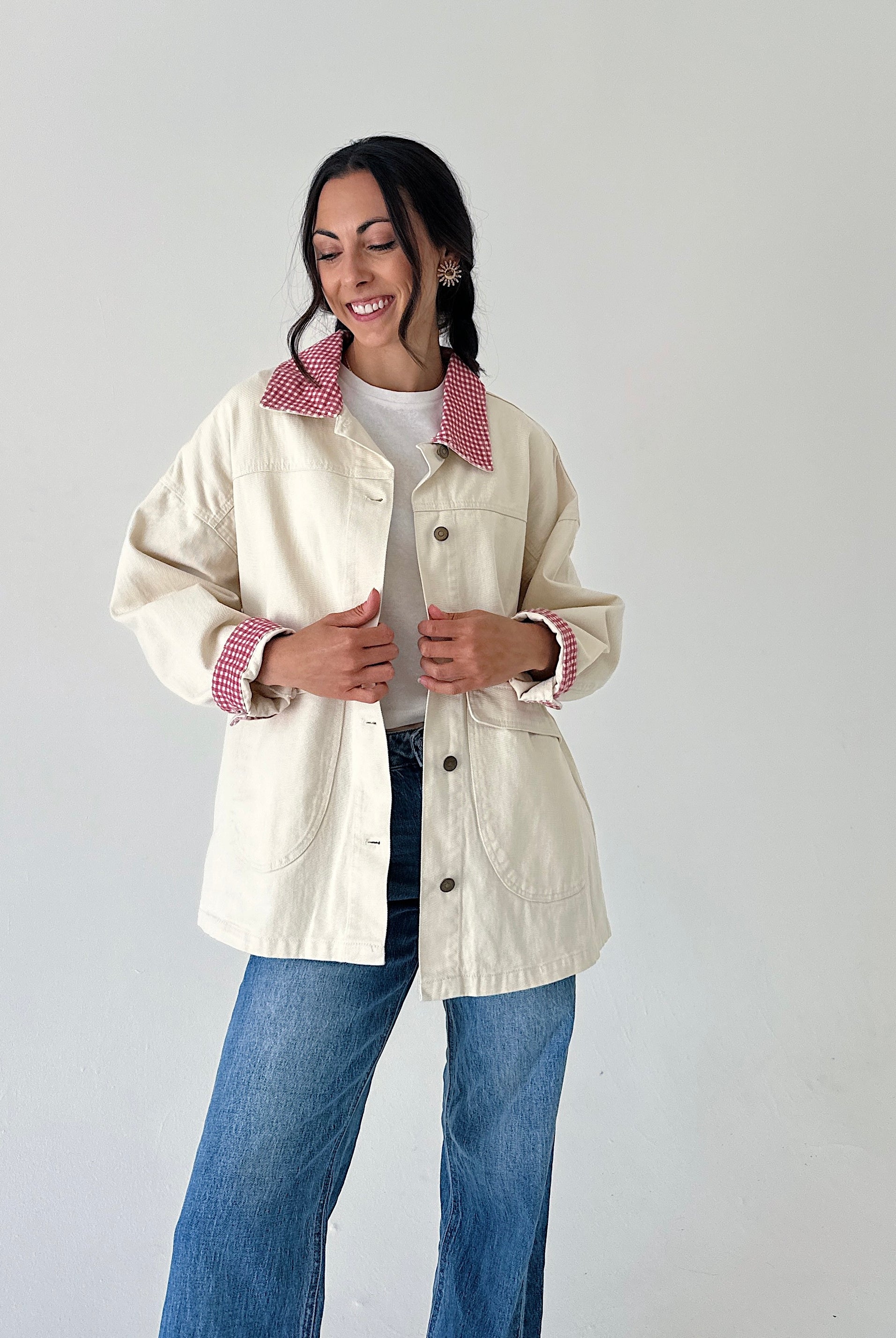 A woman modeling a cream-colored utility jacket with a red-and-white gingham patterned collar and matching gingham turned-back cuffs, paired with blue jeans against a plain light gray background.