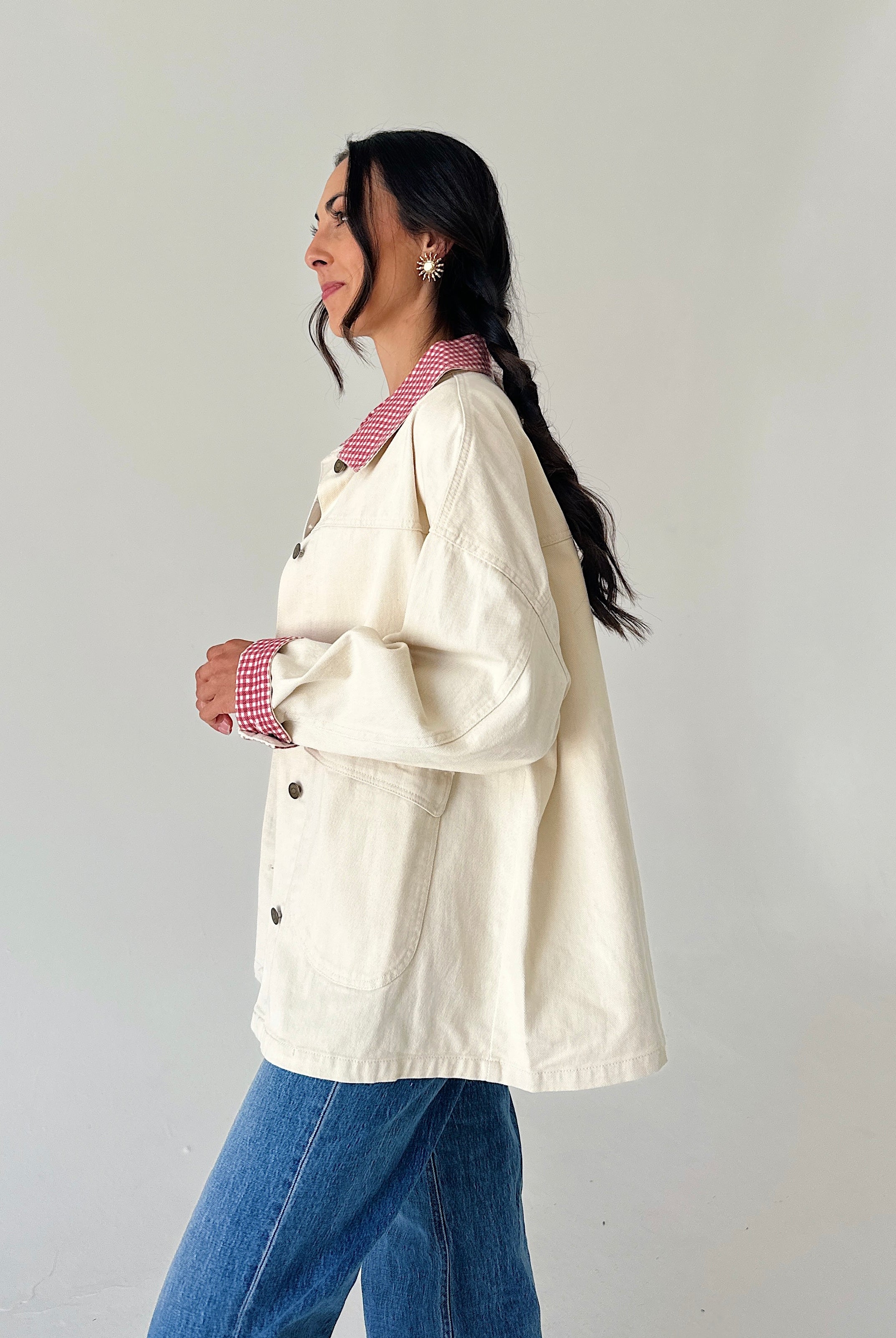 A woman wearing an oversized cream-colored utility jacket with red-and-white gingham trim on the collar and cuffs, layered over a red-and-white gingham shirt and paired with blue jeans, shown in profile against a plain light gray wall.