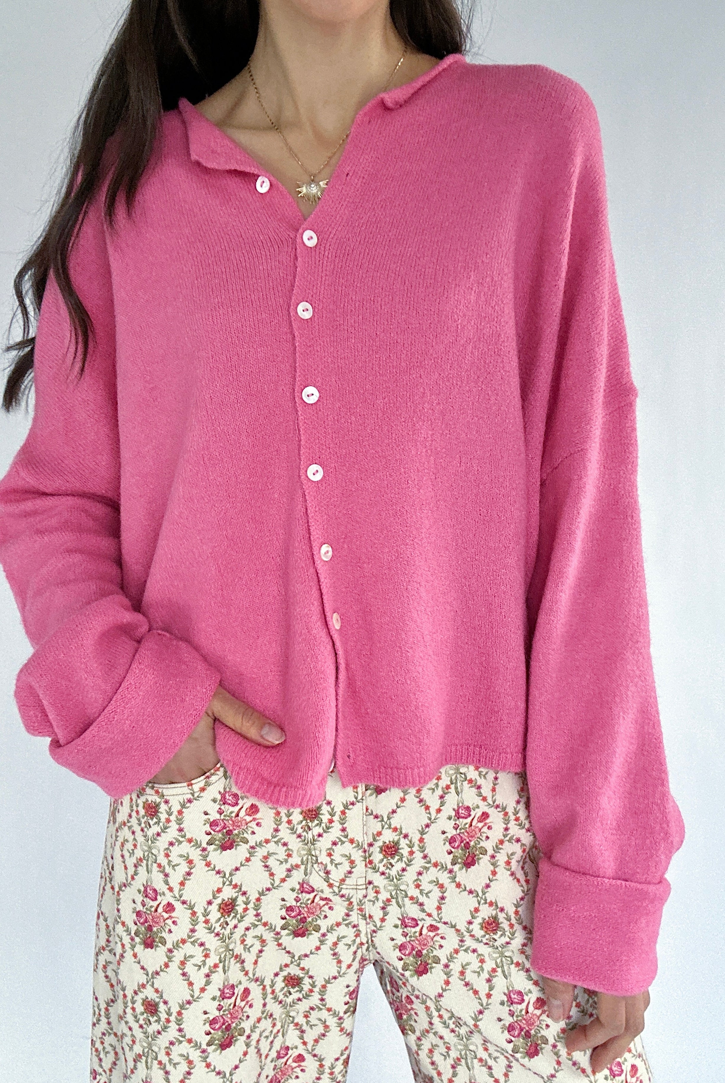 Person wearing a pink cardigan with a floral pair of pants on a plain background