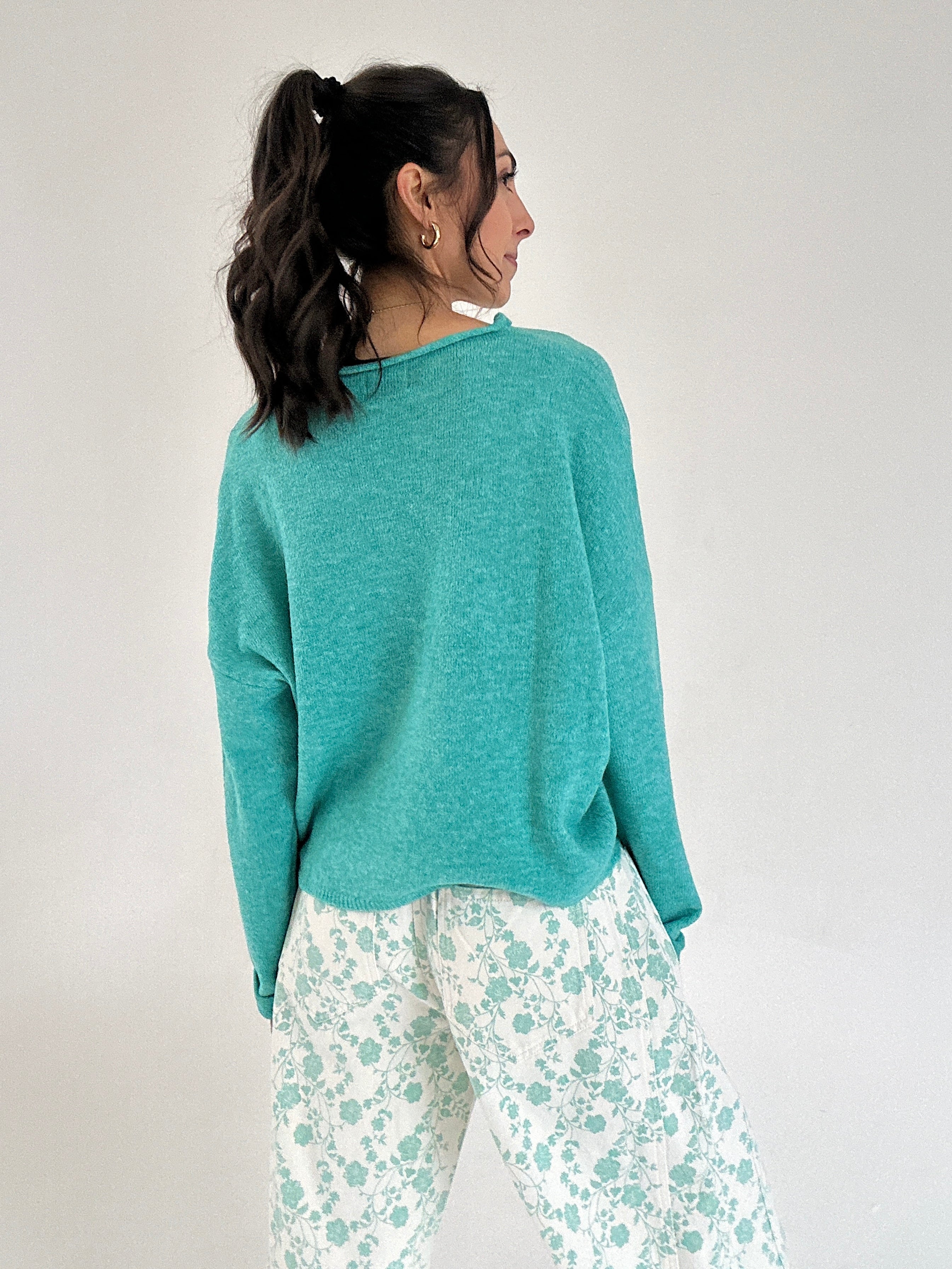 Woman wearing a teal sweater and floral pants against a plain background
