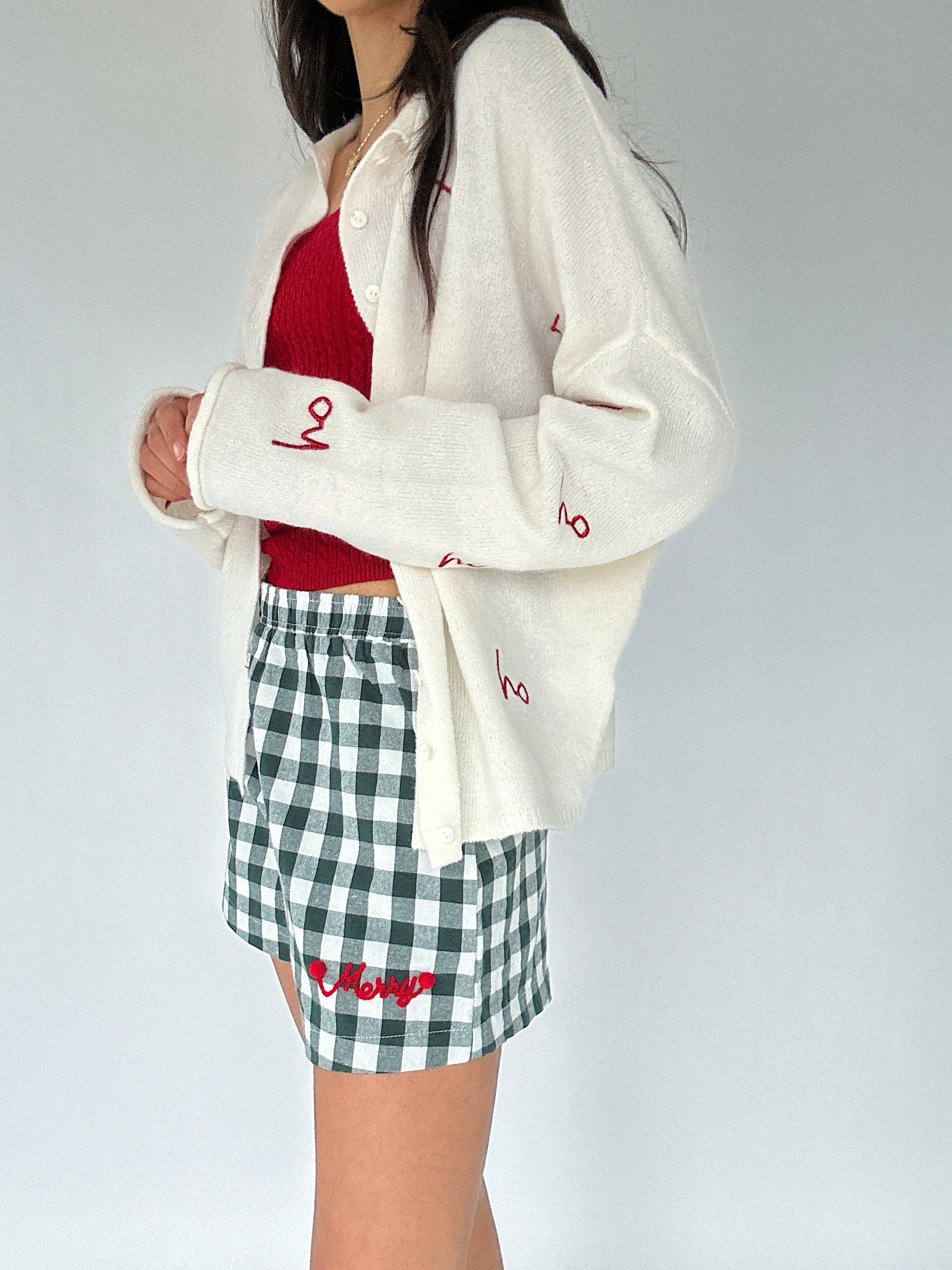 Person wearing a white cardigan with red text, red top, and green checkered shorts on a plain background