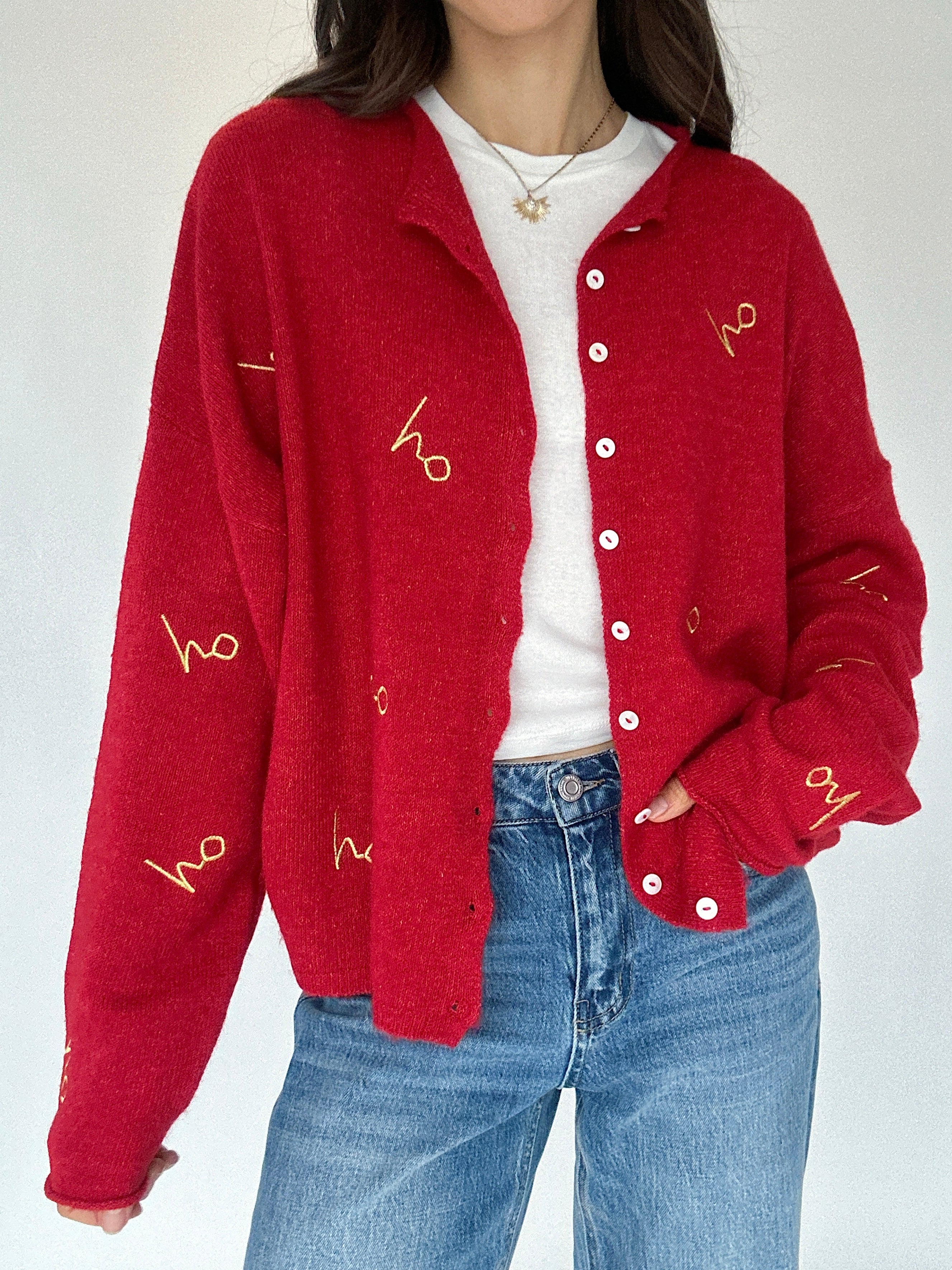 Person wearing a red cardigan with gold 'ho ho ho' text design, white shirt, and blue jeans on a white background