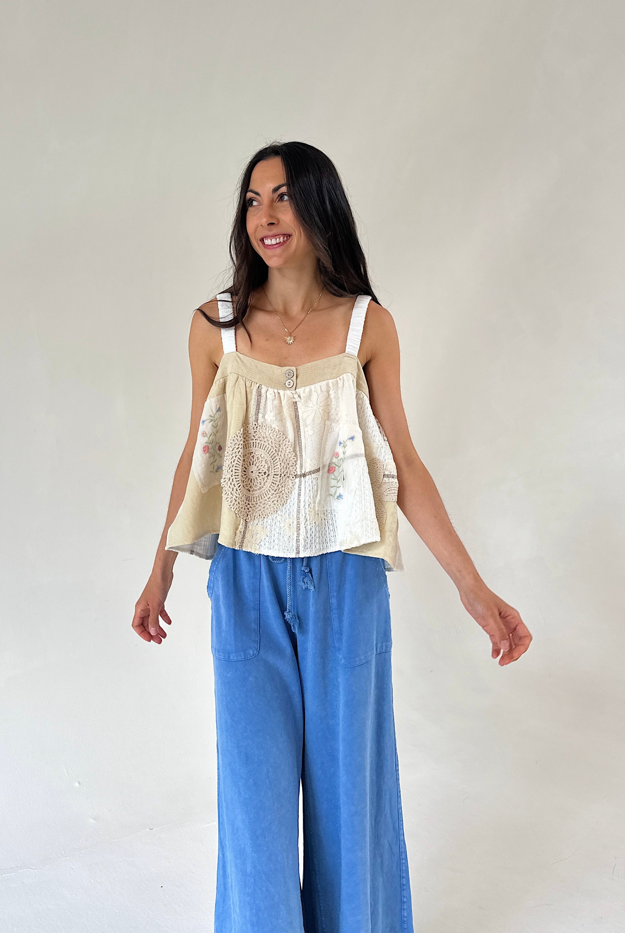 A woman modeling a cream and beige sleeveless top with intricate embroidery and a ruffled peplum hem, paired with wide-leg blue jeans, against a plain off-white studio background.